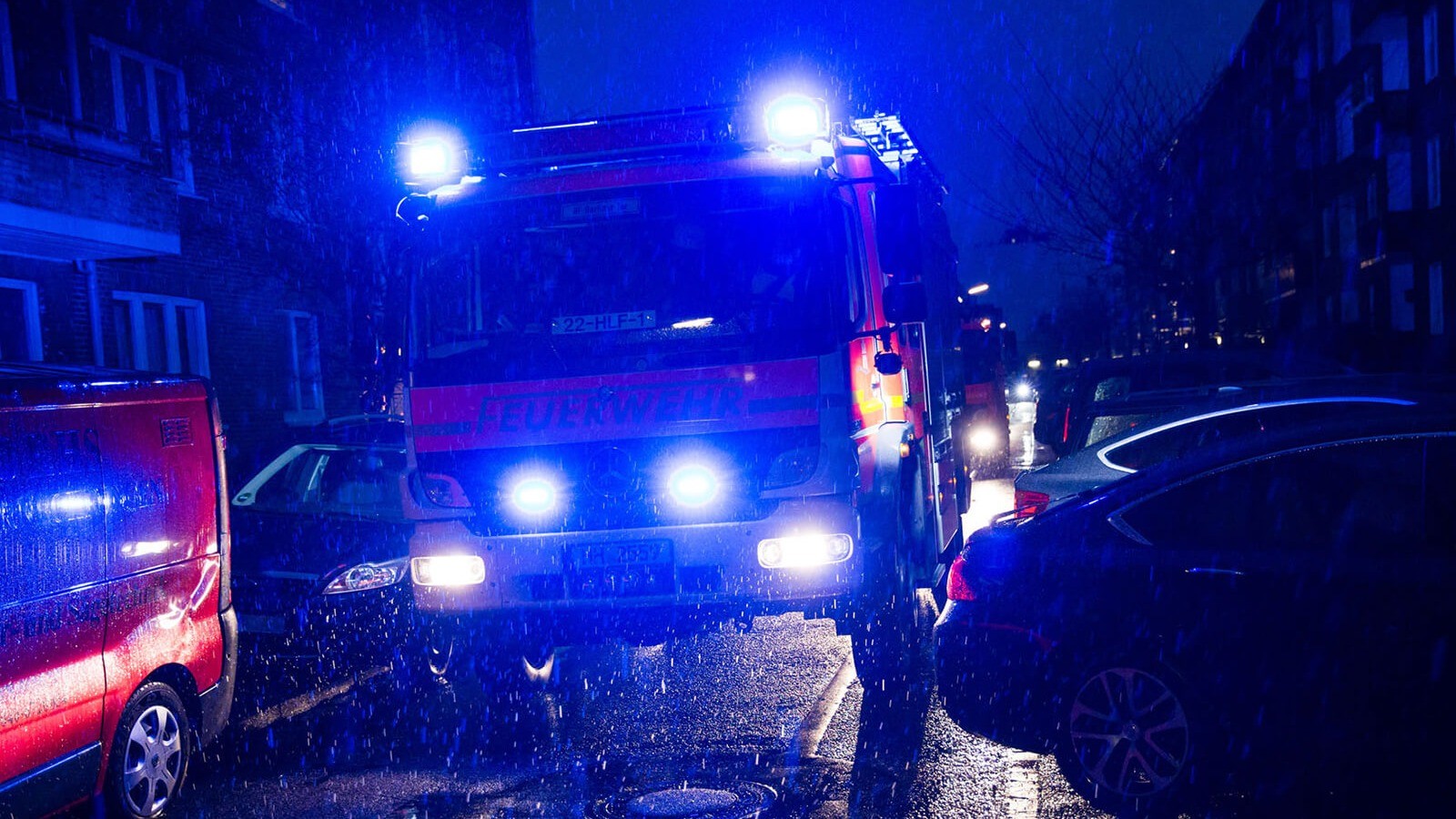 Ein Feuerwehrwagen fährt nachts mit Blaulicht durch eine sehr enge Straße, in der rechts und links Autos parken. 