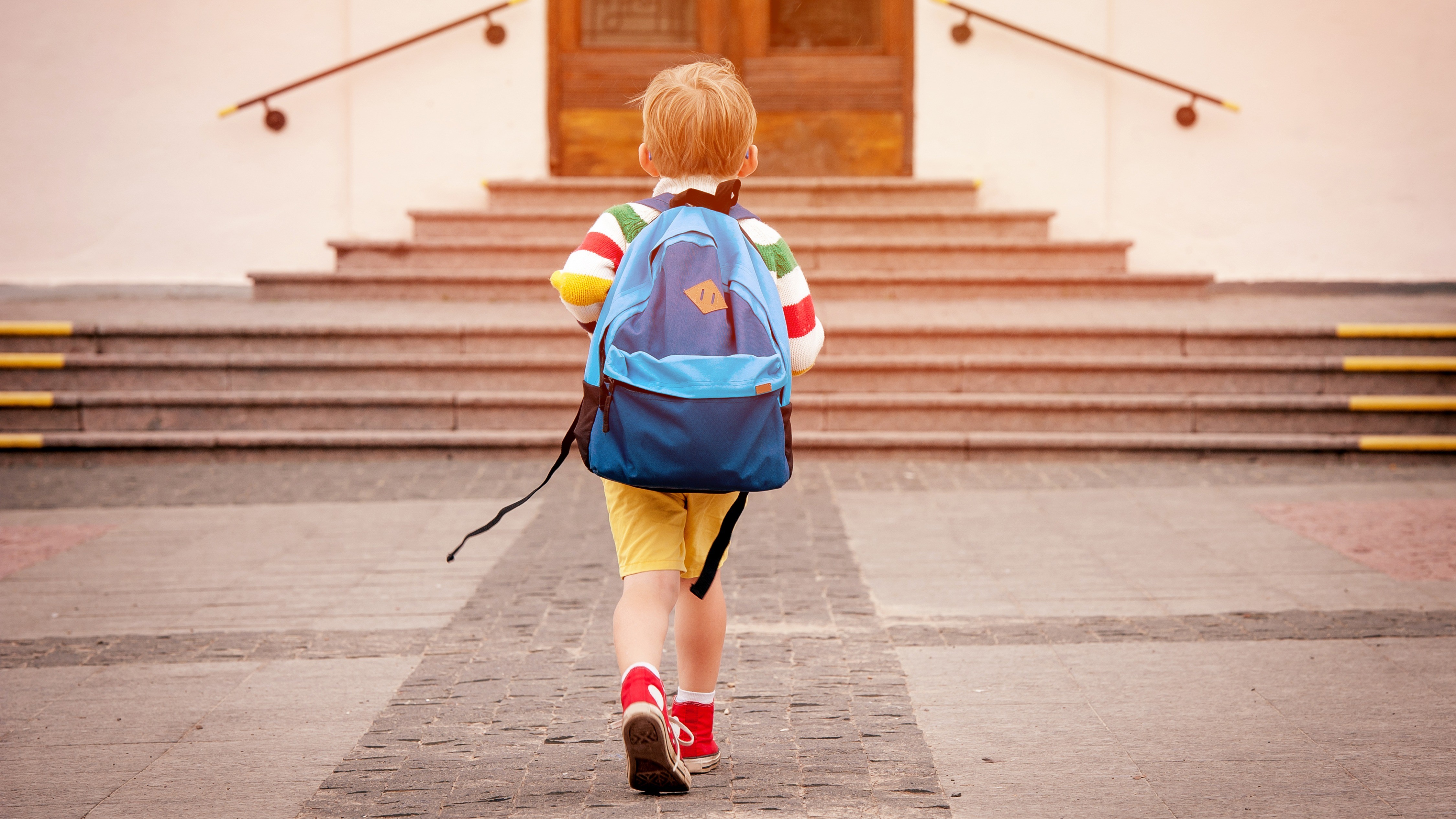 Ein Schuljunge mit einem blauen Rucksack, gelber Hose und einem bunt gestreiften Pullover läuft auf die braune Holztür eines Schulgebäudes zu.  