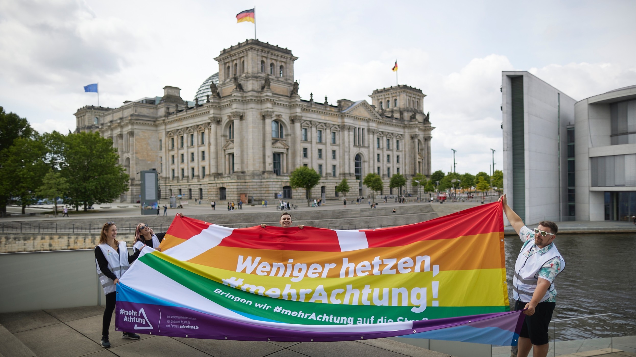Vier Menschen halten ein Stoffbanner in Regenbogenfarben. Darauf zu lesen: „Weniger hetzen, mehr Achtung!“. Im Hintergrund ist der Reichstag in Berlin zu sehen.