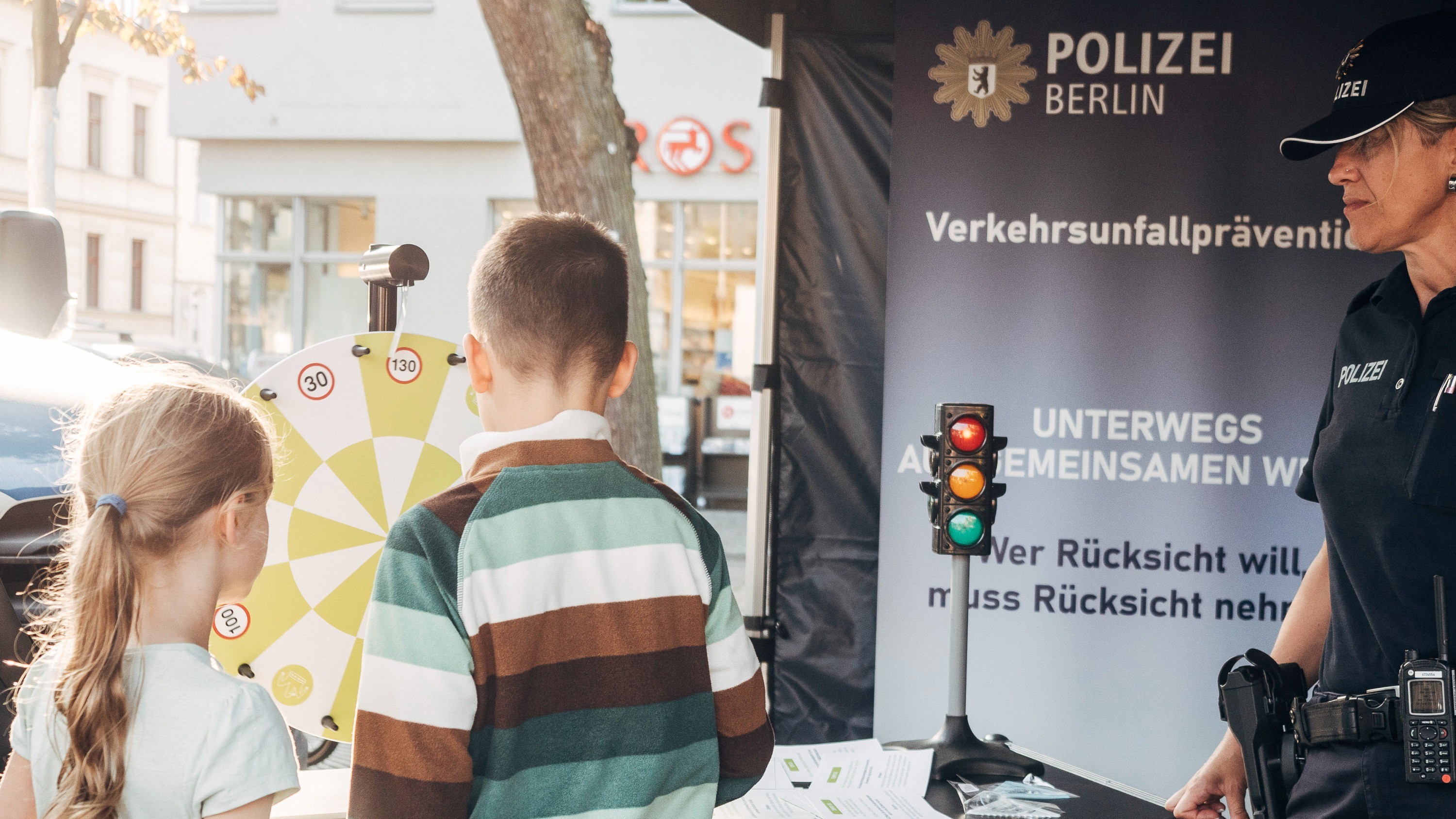 Ein Mädchen und ein Junge stehen an einem Aktionsstand und drehen an einem Glücksrad. Im Hintergrund steht ein Roll-up-Banner mit der Aufschrift „Polizei Berlin. Verkehrsunfallprävention“. Neben ihnen steht eine Polizistin.