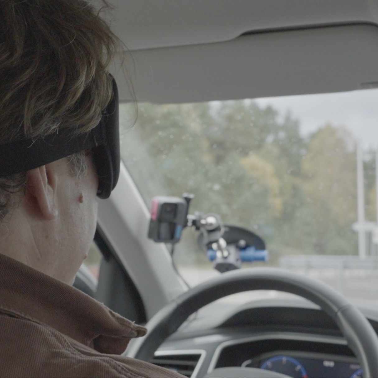 Person mit schwarzer Augenbinde auf Fahrersitz eines Pkw, Straße durch Windschutzscheibe sichtbar, Kamera am Armaturenbrett befestigt.