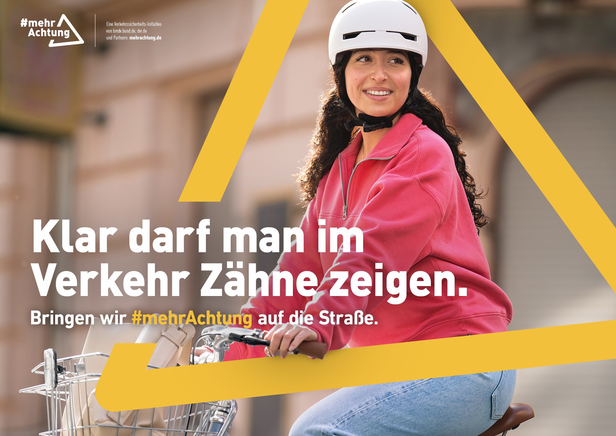 Das Bild zeigt eine Fahrradfahrerin in roter Jacke und mit weißem Helm. In einem gelben Dreieck, das grafisch über das Bild gelegt ist und an das Verkehrszeichen „Gefahrenstelle“ erinnert, steht der Text: „Klar darf man im Verkehr Zähne zeigen. Bringen wir mehr Achtung auf die Straße.“