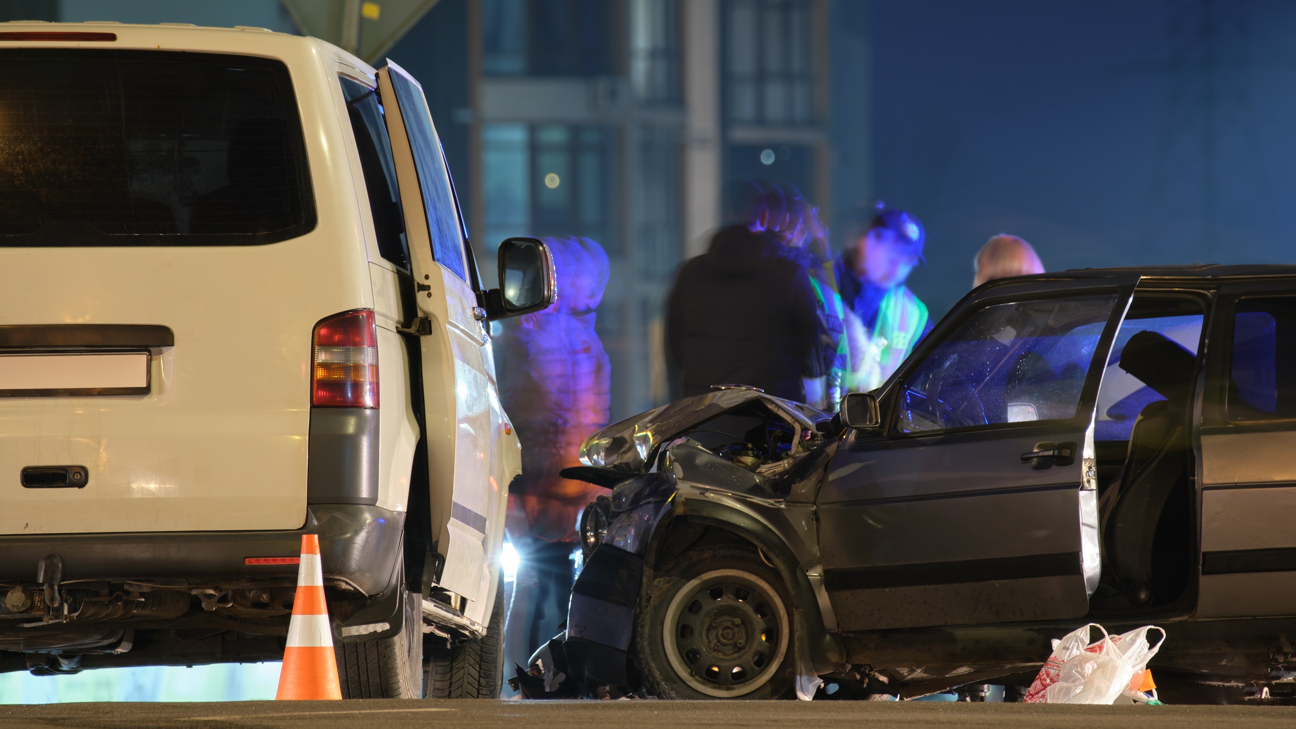 Bild Verkehrsunfall-Opferhilfe 16:9 Zwei Autos nachts nach einem Verkehrsunfall. Das Auto rechts im Bild ist stark beschädigt. Im Hintergrund sind Silhouetten verschiedener Personen zu erkennen.