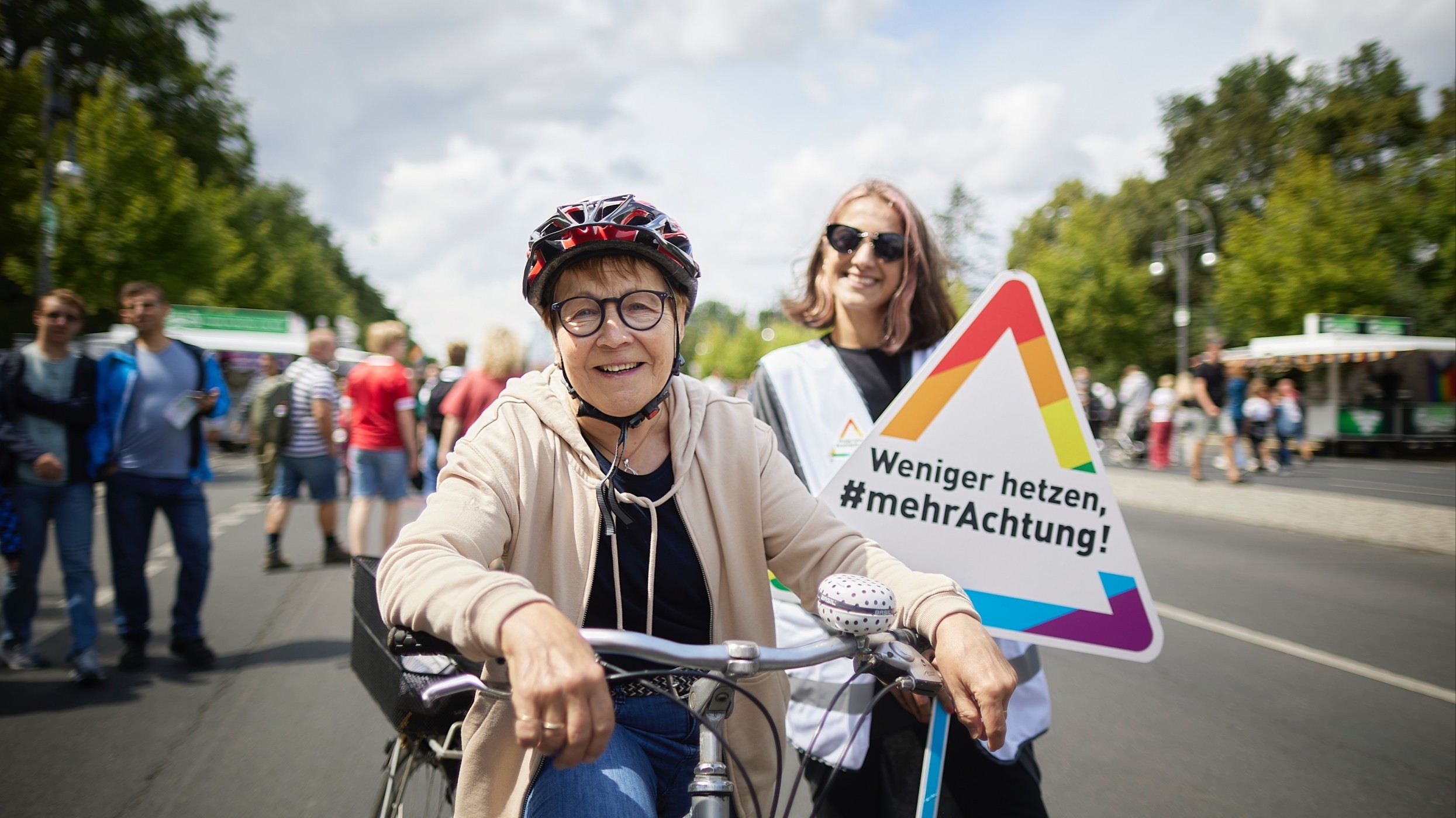 Eine Radfahrerin steht mit ihrem Fahrrad auf einer abgesperrten Straße. Neben ihr ist eine junge Frau zu sehen. Sie hält ein dreieckiges Schild in Regenbogenfarben mit der Aufschrift „Weniger hetzen, mehr Achtung!“.