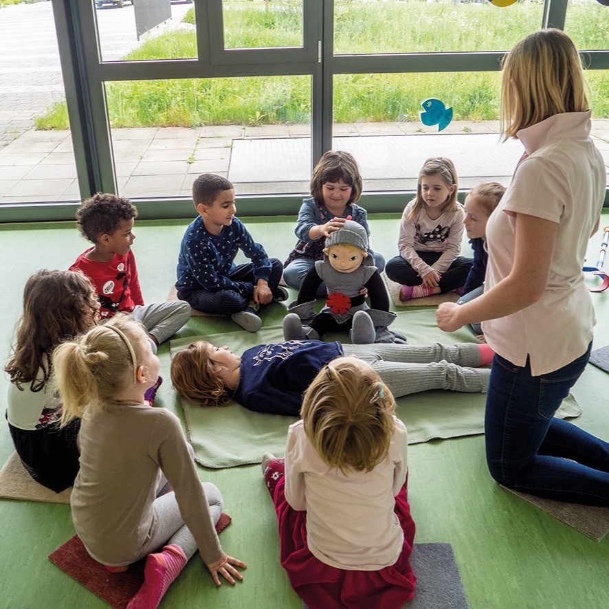 mA von klein auf Dank Partner Gruppe Kinder Eine Gruppe Kinder sitzt auf grünem Boden im Kreis um die Handpuppe „Ritter Björn“. Ein Kind hält die Puppe, eine Frau kniet daneben.