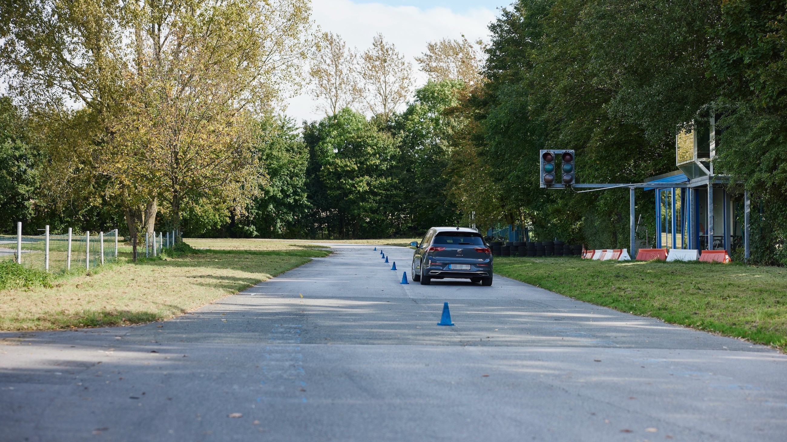 Fahrsicherheitstraining Slalom Ein Auto fährt auf einer Straße mit blauen Verkehrskegeln, die eine Spur markieren. Rechts ist eine ausgeschaltete Ampel zu sehen. Die Umgebung ist von Bäumen gesäumt.