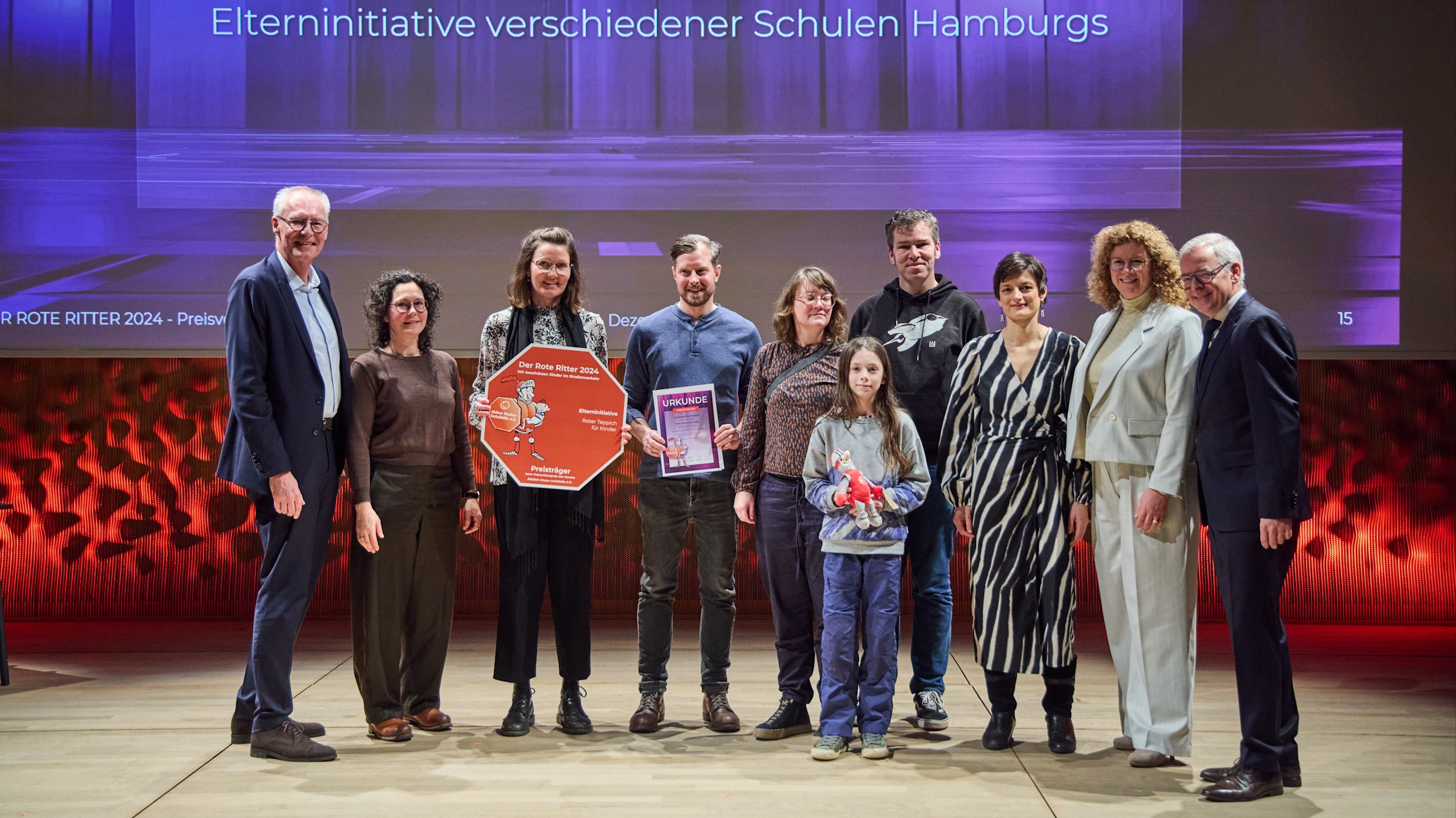 16:9 Roter Ritter Preisverleihung Auf einer Bühne steht eine Gruppe von Frauen, Männern und einem Kind. Sie halten eine Urkunde und einen Preis in den Händen. Es ist das Siegerprojekt: "Roter Teppich für Kinder".