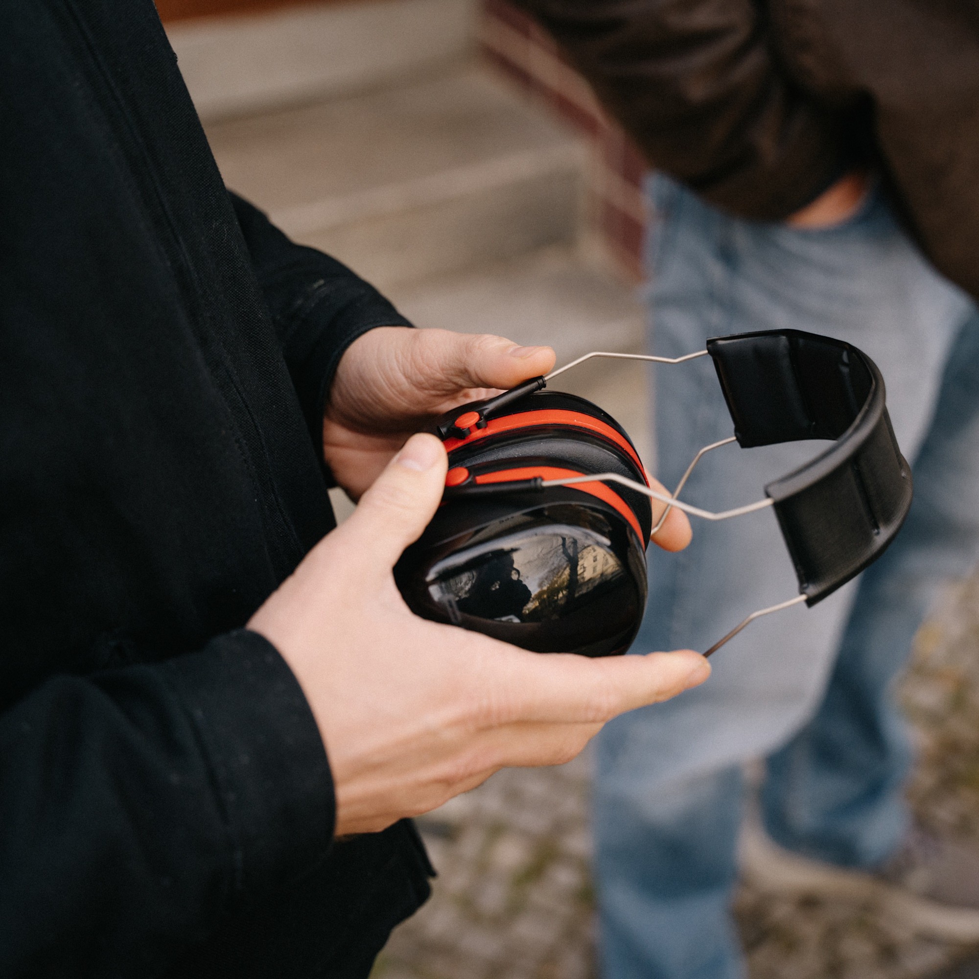 Schwerhörigkeit Kapselhörschutz 1:1 Nahaufnahme der Hände einer Person, die einen schwarz-roten Kapselgehörschutz halten. Im unscharfen Hintergrund steht eine weitere Person in Jeans und brauner Jacke.