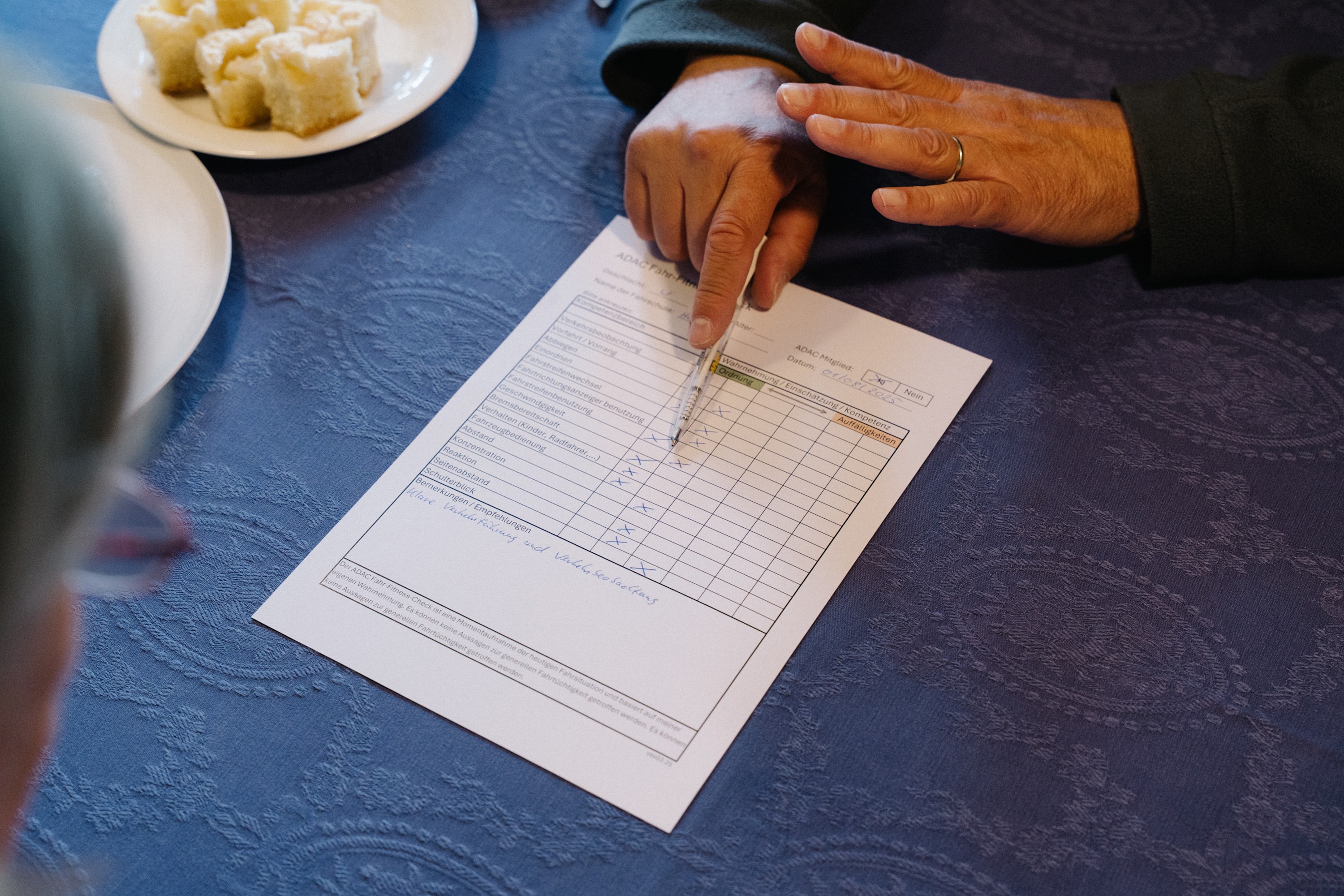 Nahaufnahme eines Formulars mit dem Titel „ADAC Fahr-Fitness-Check“ auf einem blauen Tischtuch. Die Hand eines Mannes zeigt mit einem Stift auf eine angekreuzte Checkliste auf dem Dokument.
