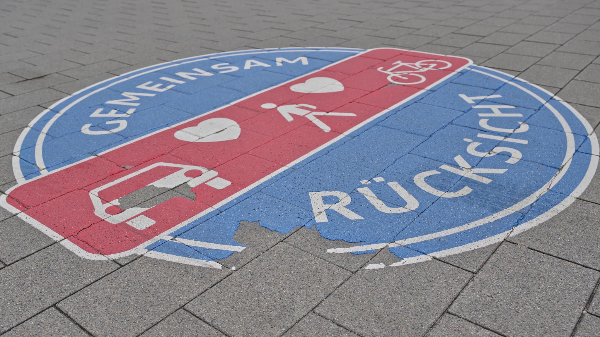 Eine runde Bodenmarkierung auf grauem Pflasterstein, die einem Verkehrsschild ähnelt und die Aufschrift „Gemeinsam” und „Rücksicht” trägt. In der Mitte befindet sich ein weiteres rotes Symbol, das ein Auto, ein Fahrrad und einen Fußgänger gemeinsam darstellt und von zwei Herzen umgeben ist.