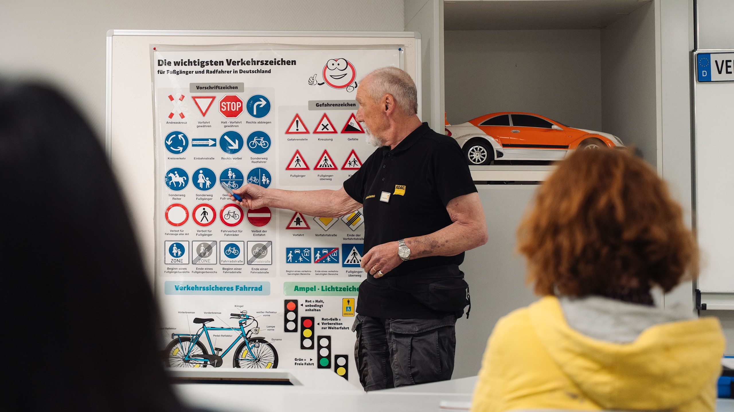 Verkehrstrainer Wolfgang Weschke steht in einer Verkehrsschule vor den Teilnehmenden und zeigt auf ein Plakat, auf dem „Die wichtigsten Verkehrszeichen“ abgebildet sind.
