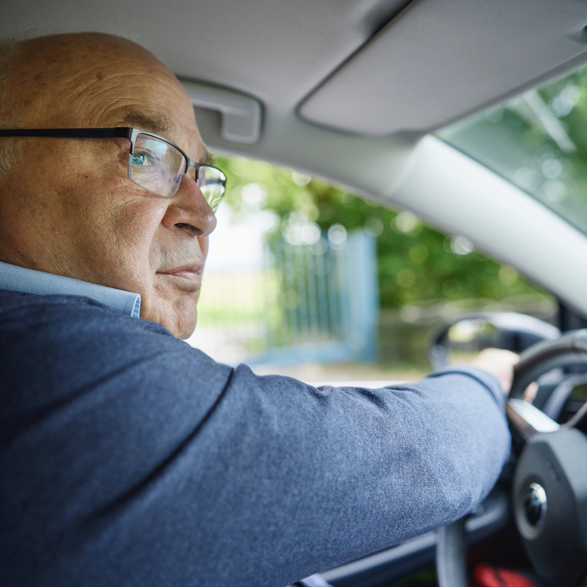 Fahrsicherheitstraining Bernd Henne Ein älterer Mann mit Brille sitzt am Steuer eines Autos und schaut in den rechten Seitenspiegel. Die Hand liegt auf dem Lenkrad, und durch das Fenster sind Bäume zu sehen.