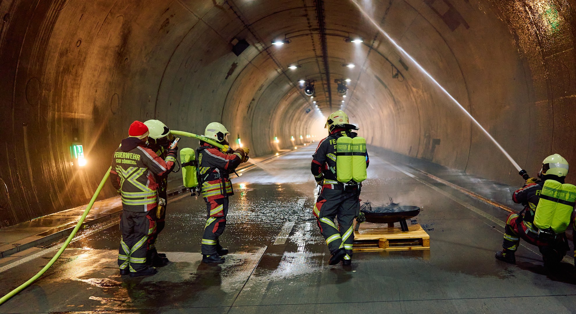 Feuerwehrleute löschen den simulierten Brandherd. Zeitgleich besprüht ein Kamerad die Tunnelwand mit Wasser und kühlt sie damit.