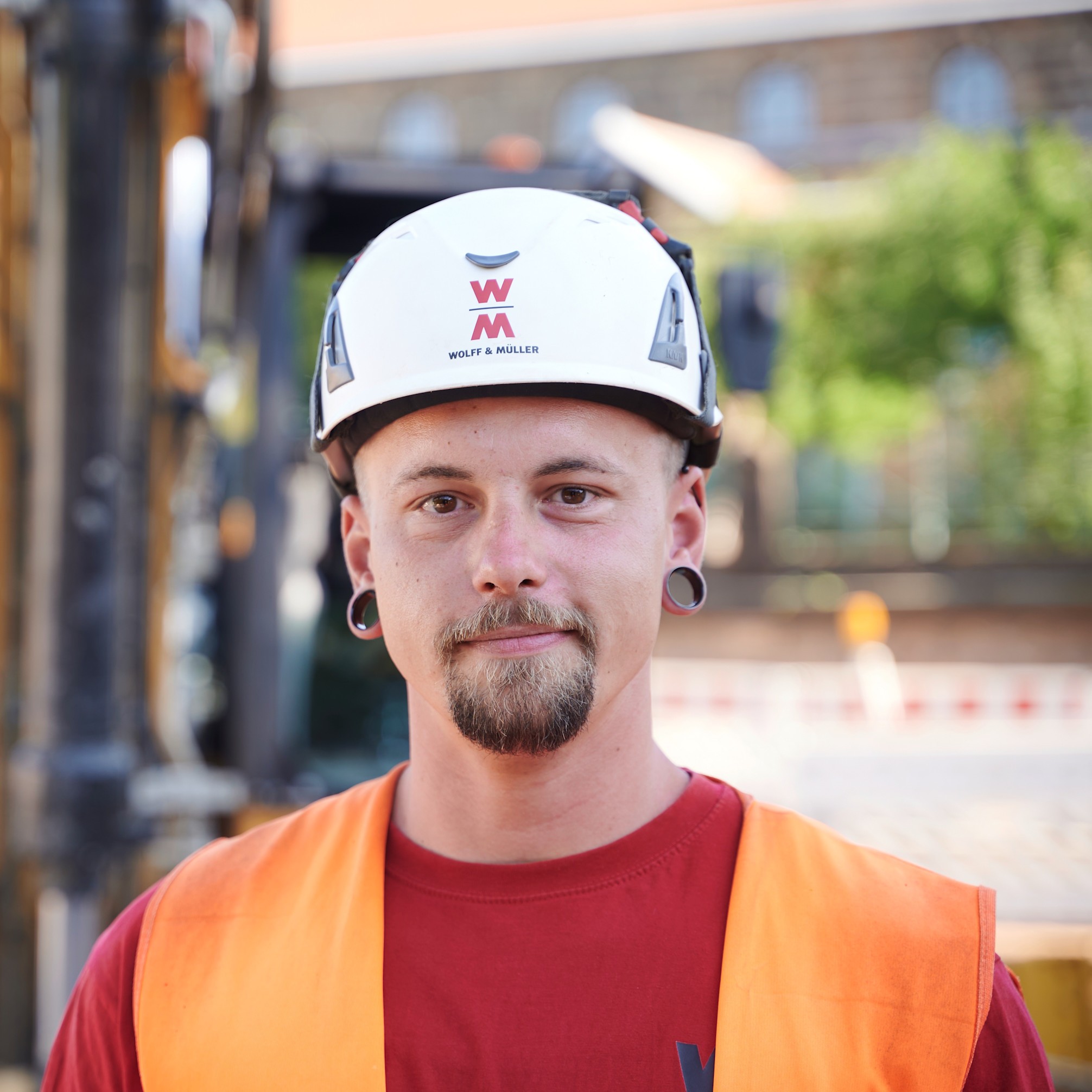 Ein Porträt eines jungen Mannes mit Bart, der eine orangefarbene Warnweste und einen weißen Helm trägt. Im Hintergrund ist ein Bagger zu sehen.