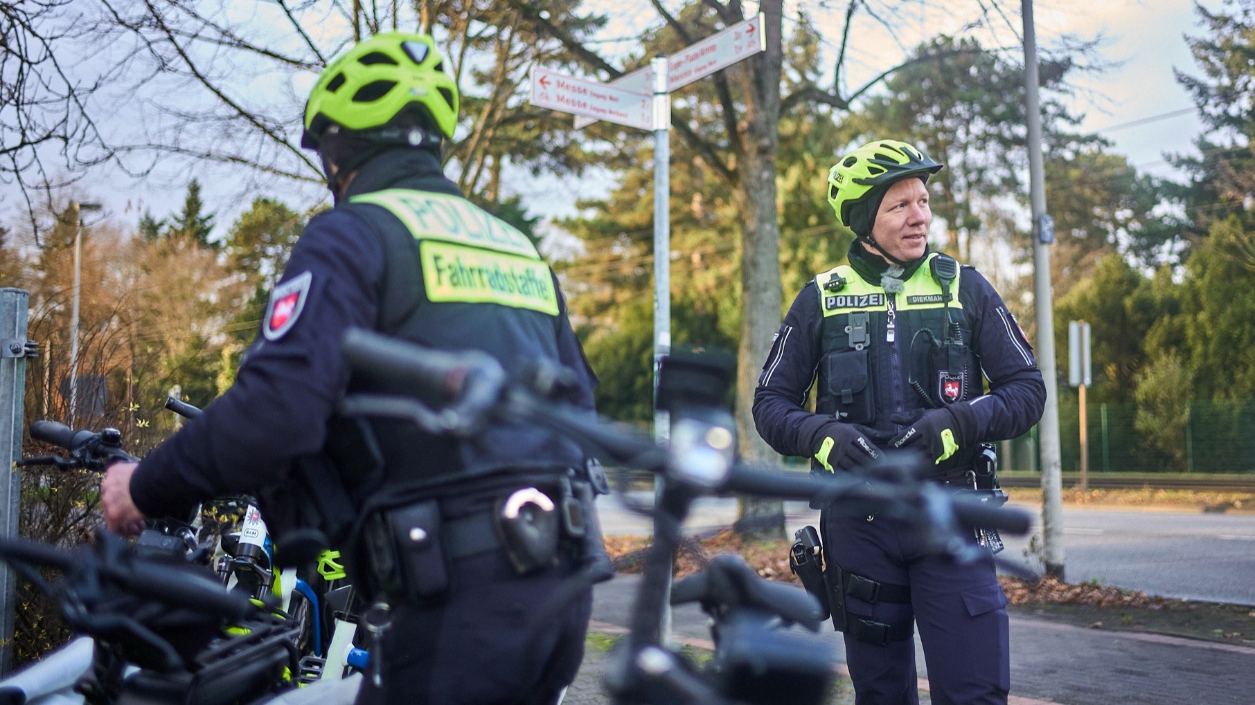 Fabian Koester Polizei Fahrradstaffel Bilder Polizei 16:9 Zwei Polizisten in dunkelblauer Uniform stehen neben ihren Fahrrädern auf einem Weg. Beide tragen leuchtend gelbe Fahrradhelme.