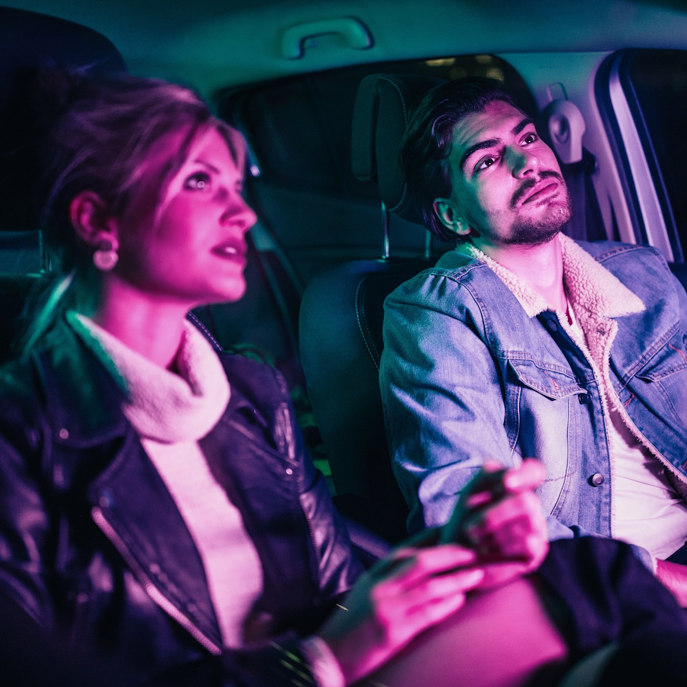 Bild Im Taxi 2 (1:1) Zwei junge Leute sitzen auf der Rückbank eines Autos und blicken in verschiedene Richtungen. Das pink-lila Licht im Wageninneren beleuchtet ihre Gesichter.