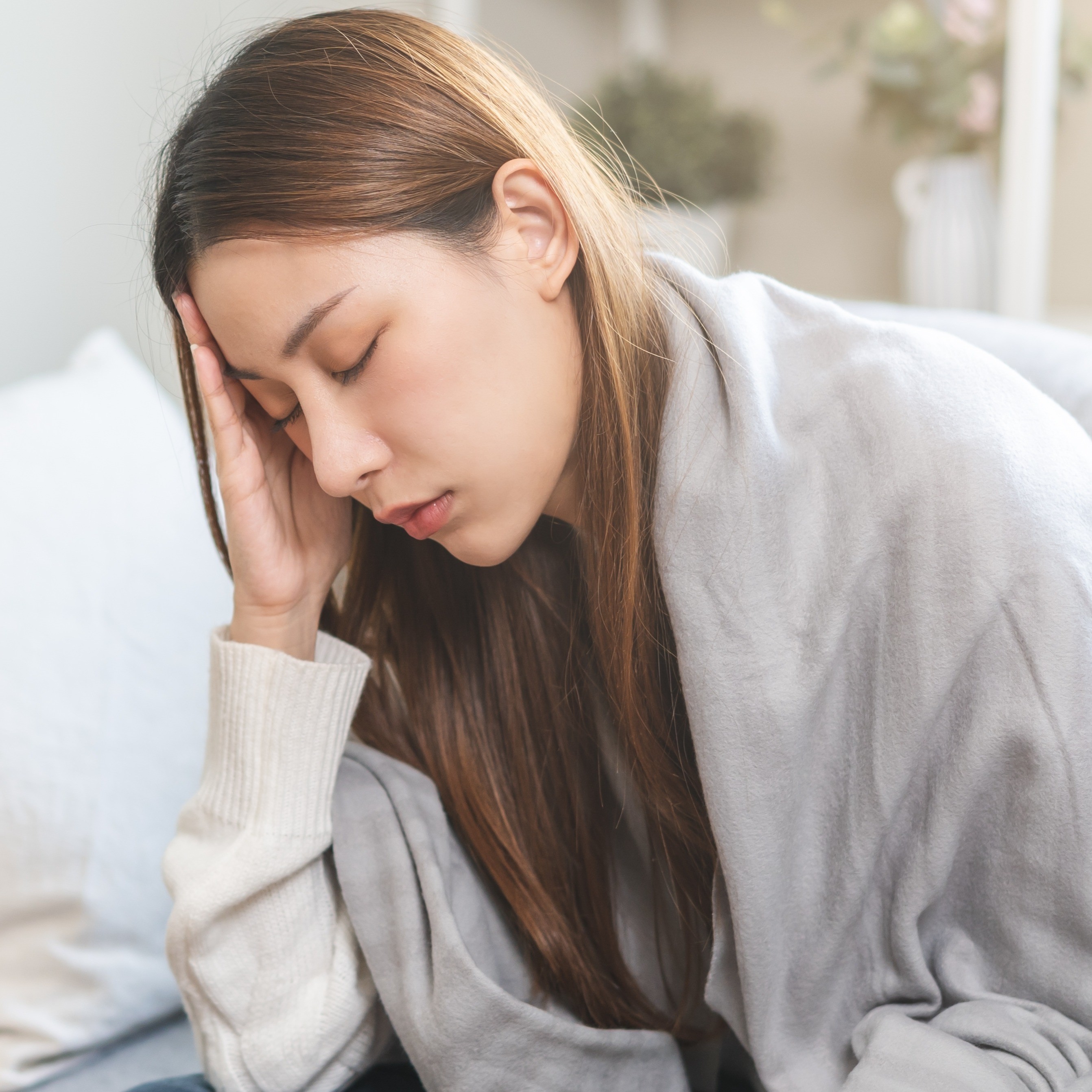 Eine junge Frau hält sich mit geschlossenen Augen die Hand an den Kopf. Sie fühlt sich unwohl, hat Kopfschmerzen und liegt zugedeckt auf einem Sofa.