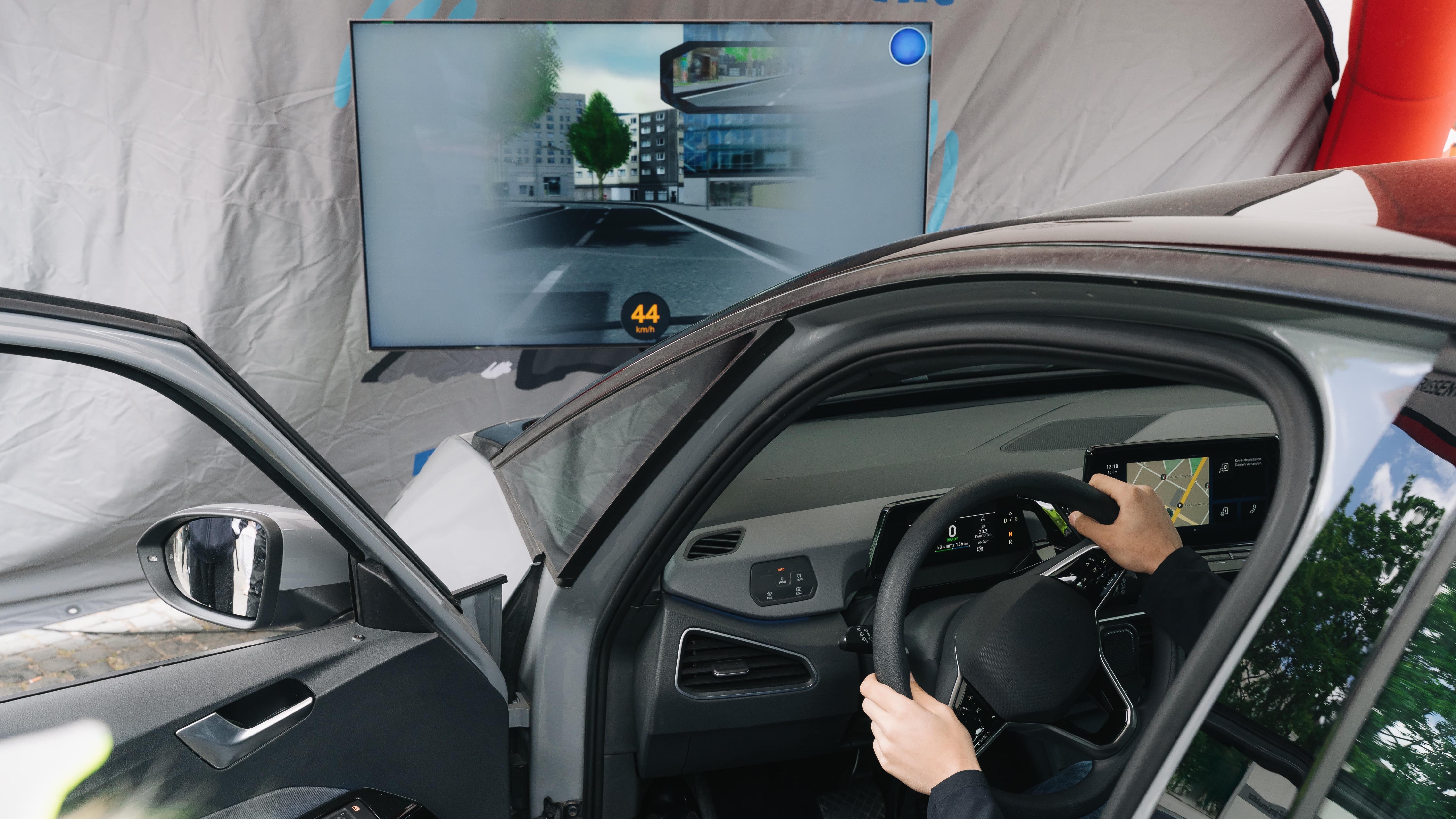 Ein Pkw-Fahrsimulator mit geöffneter Tür zeigt eine Testperson am Steuer. Auf dem Bildschirm simuliert eine virtuelle Stadt die Auswirkungen von Alkohol am Steuer – inklusive Tunnelblick. Im Hintergrund ein Banner zur Wirkung von Alkohol, im Innenraum sind Display und Bedienelemente sichtbar.