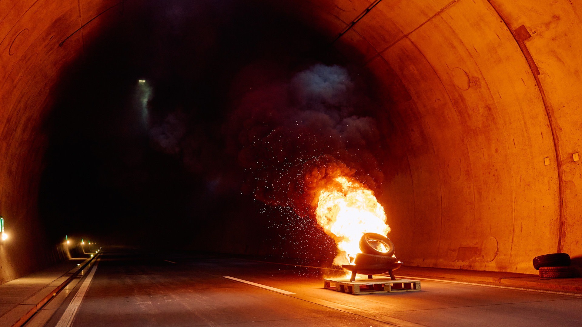 Ein intensives Feuer aus brennenden alten Reifen auf einer Holzpalette im Zentrum eines dunklen Straßentunnels. Der Schein des Feuers beleuchtet die Tunnelwände. Im Hintergrund ist dichter, dunkler Rauch zu sehen. 