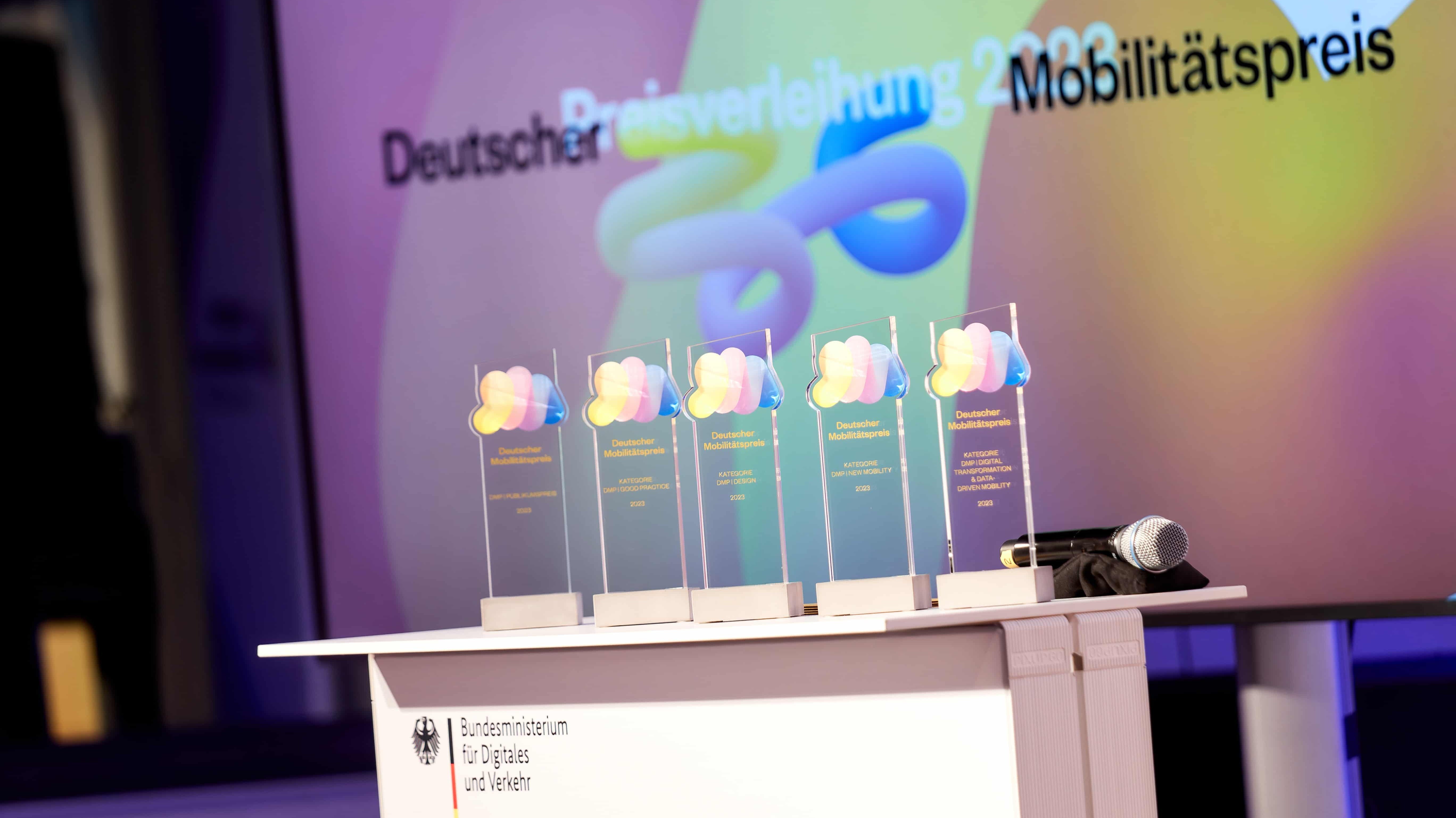 Dt_Mobilitaetspreis_Trophaen Auf einem weißen Tisch stehen fünf durchsichtige Trophäen. Zu sehen sind die Auszeichnungen des Deutschen Mobilitätspreises 2023.