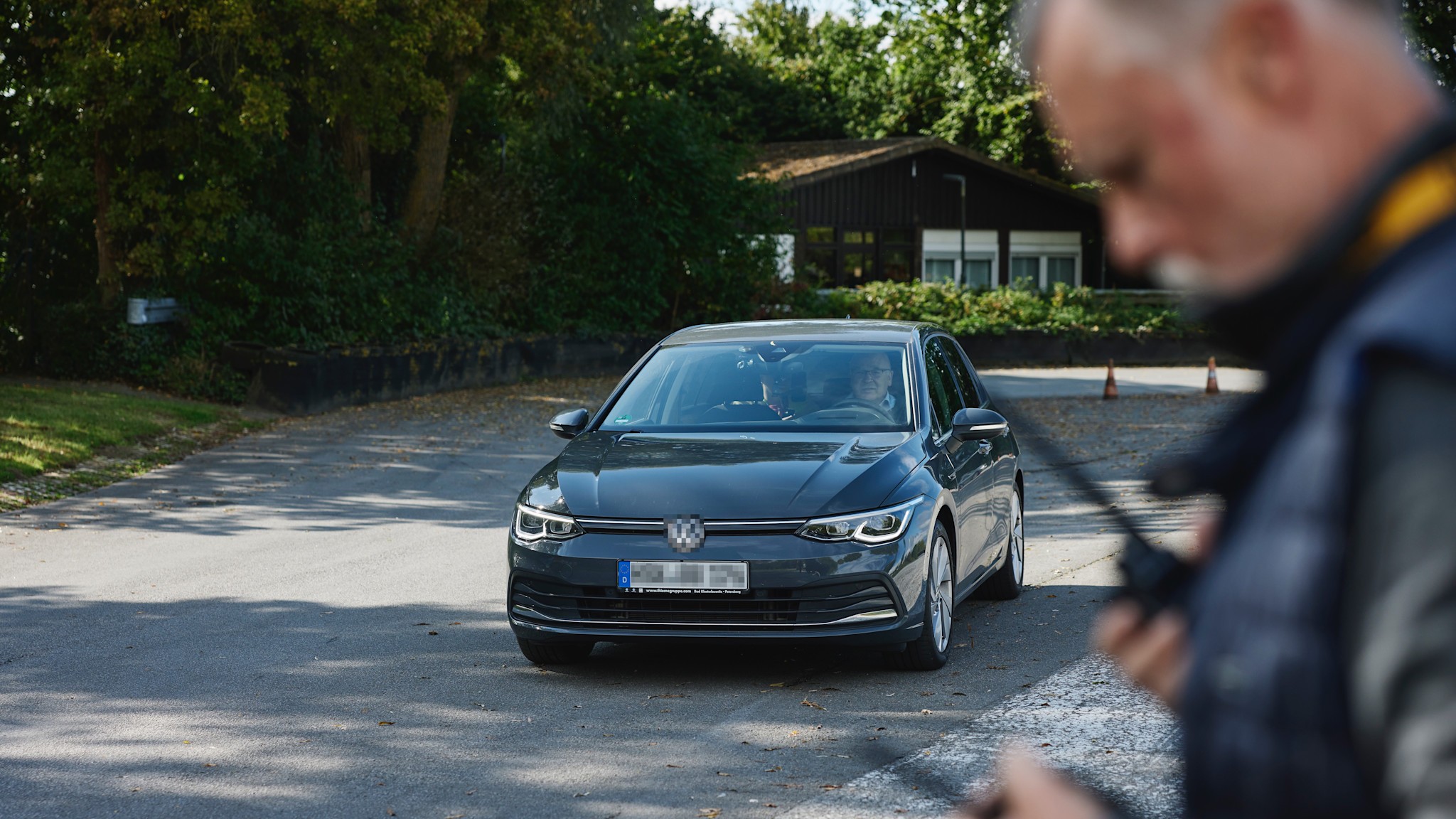 Fahrsicherheitstraining Strasse und Funkgeräte Ein graues Auto steht auf einer Straße, umgeben von Bäumen und einem Gebäude im Hintergrund. Im Vordergrund hält eine Person zwei Funkgeräte.