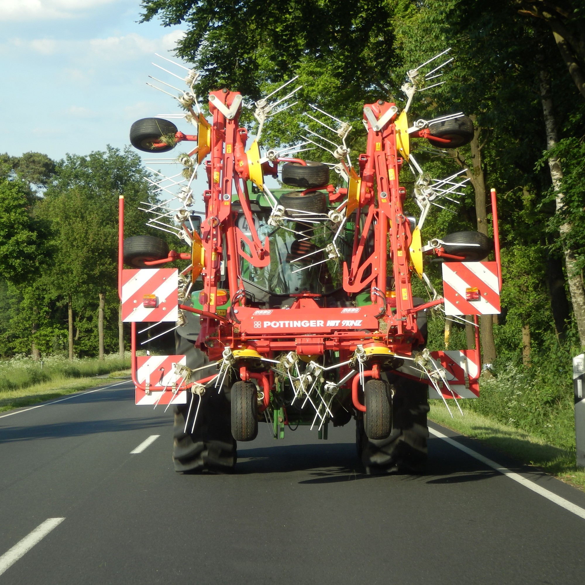 Traktor von hinten 1:1 Auf einer Landstraße ist ein Traktor mit einem großen, roten Anbaugerät unterwegs. Am Heck des Geräts sind rot-weiß gestreifte Warntafeln zu sehen.