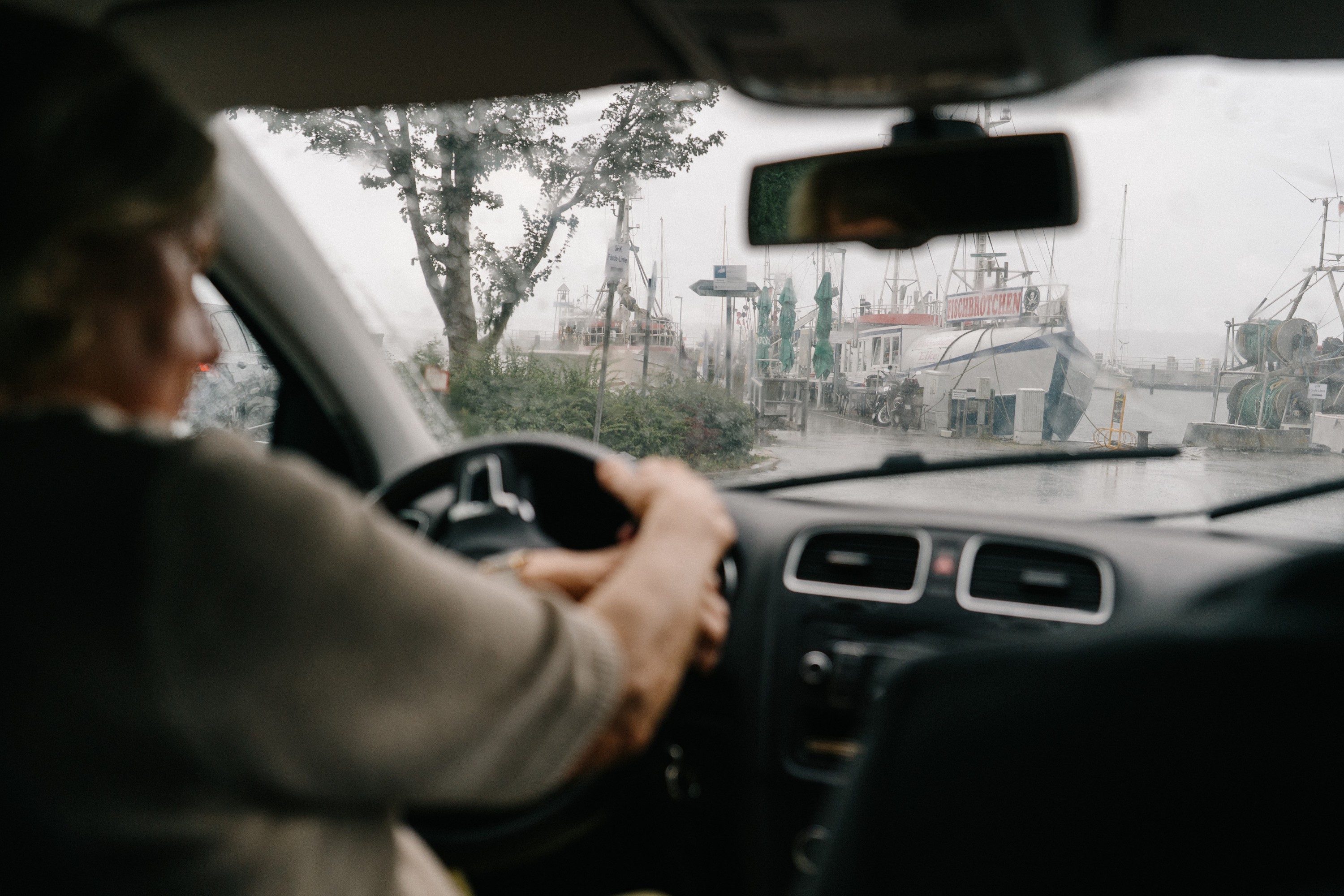 Blick aus dem Auto durch eine regennasse Windschutzscheibe: Hände am Lenkrad, im Hintergrund unscharf ein Hafen mit Schiffen unter bewölktem Himmel.