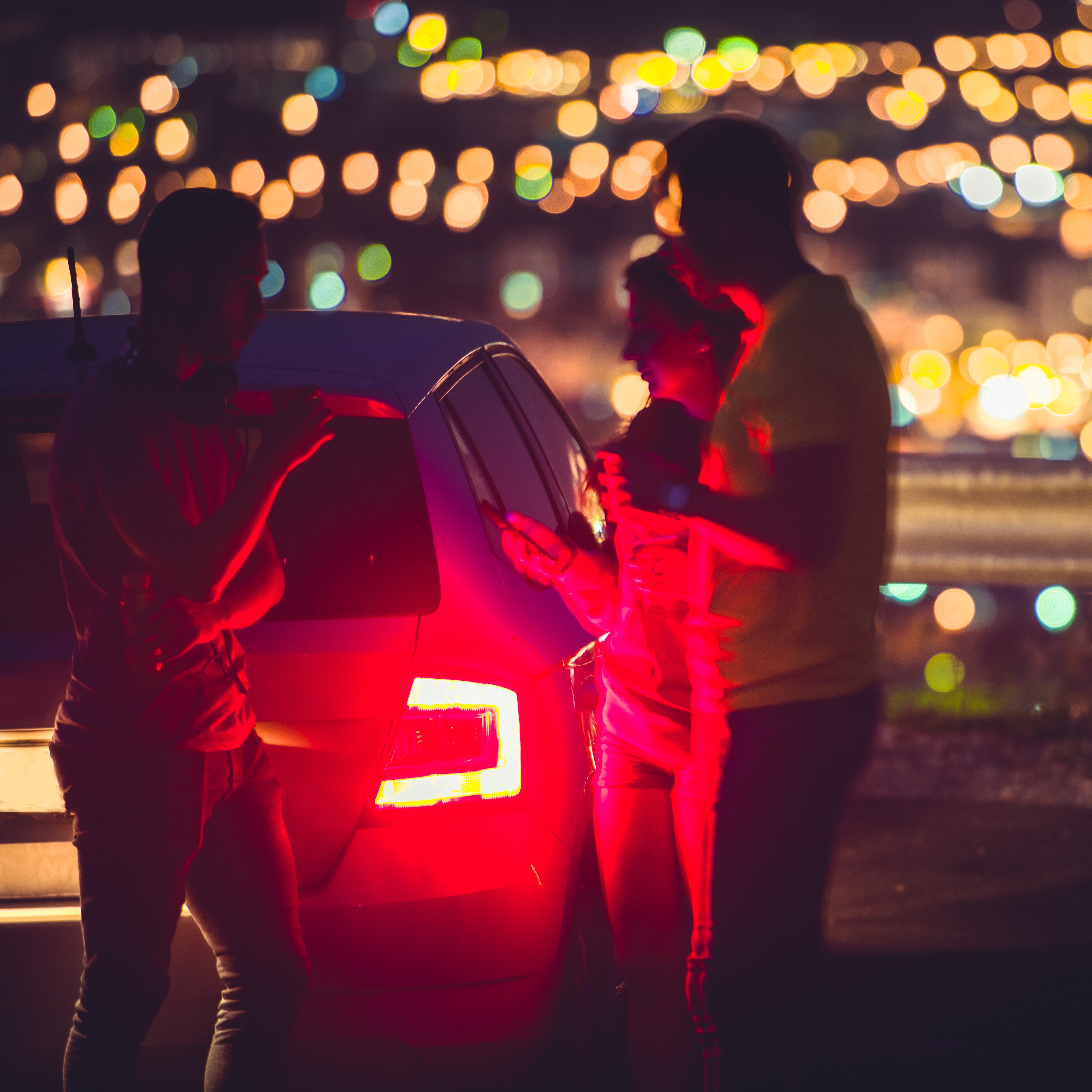 Drei Personen stehen nachts neben einem Auto, im Hintergrund sind Lichter einer Stadt zu sehen.