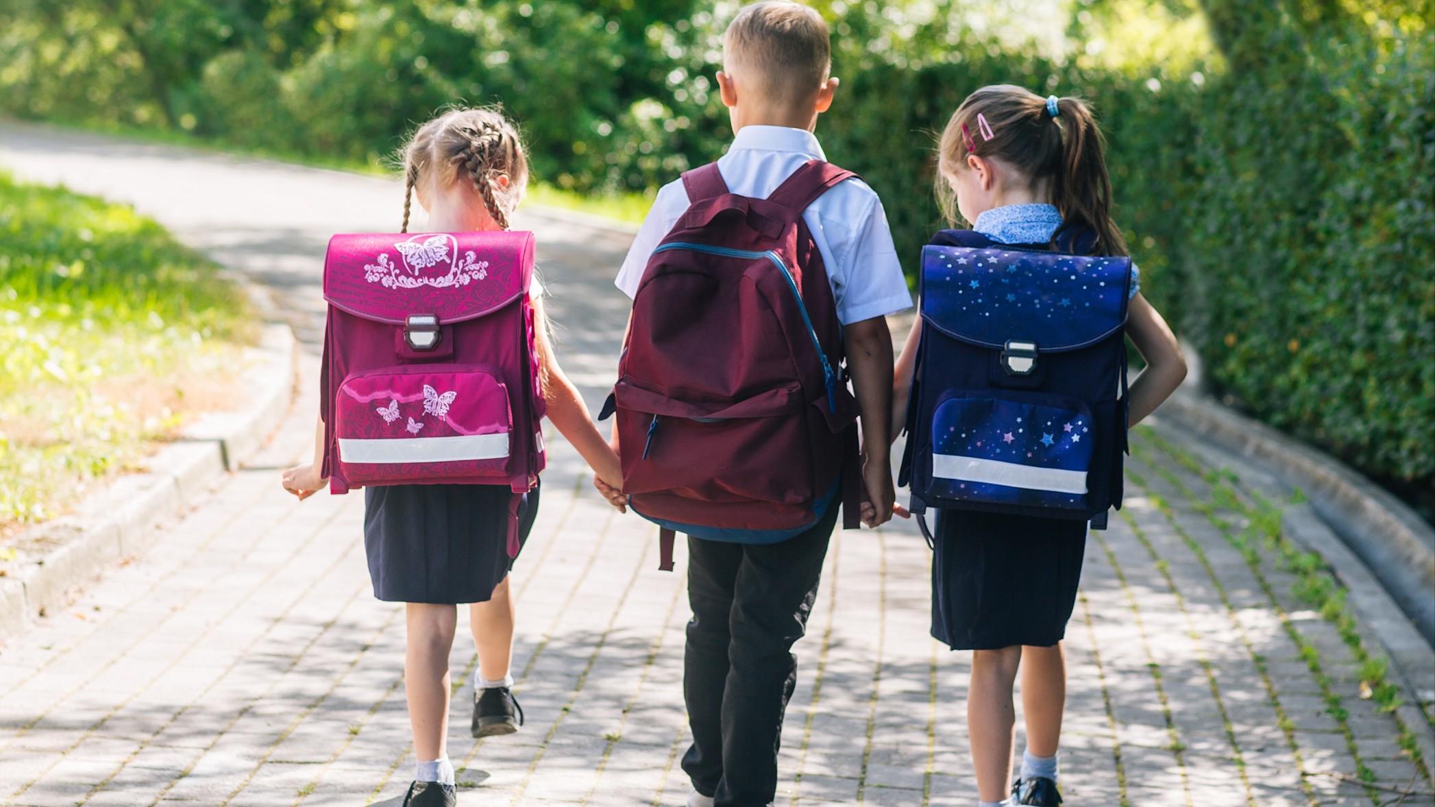 Drei Kinder laufen auf einem Steinweg. Zwei der Kinder tragen Rucksäcke mit Reflektoren. 