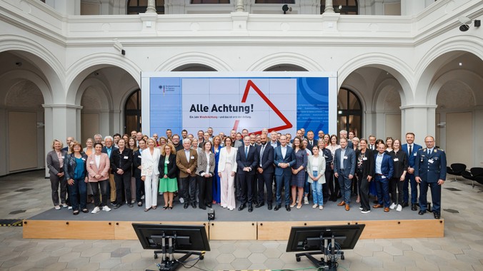 Partner Gruppenfoto Eine Gruppe von Menschen steht in einer Halle auf einer Bühne. Dahinter ist ein großes Poster mit der Aufschrift: Alle Achtung. Es sind die Partner von #mehrAchtung.
