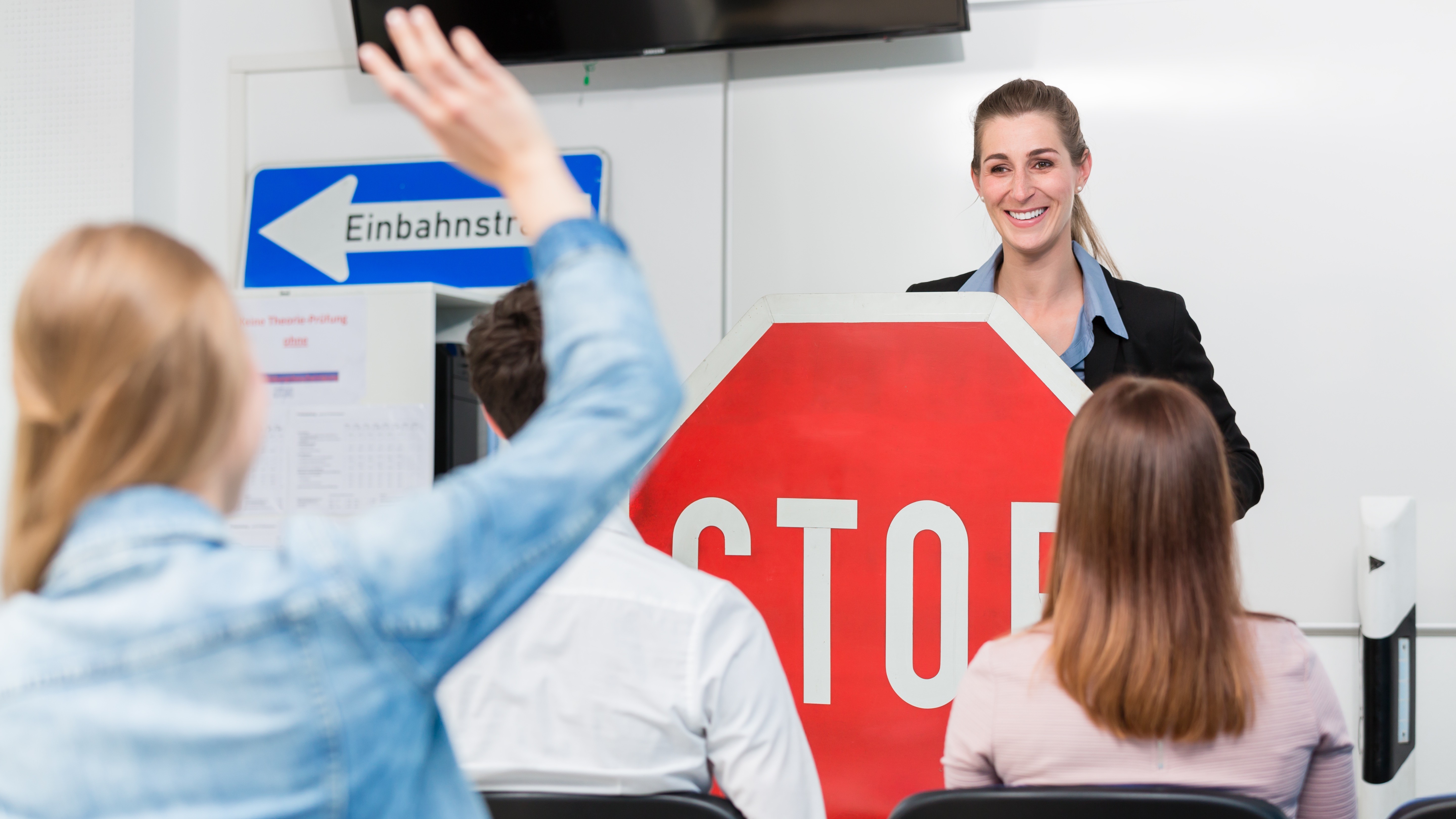 Führerscheinprüfung – Fahrschule 16:9 Eine Fahrlehrerin steht vor einer Gruppe von Fahrschülerinnen und Fahrschülern, die ein Stoppschild halten. Eine Fahrschülerin hat die Hand gehoben, um eine Frage zu stellen. Im Hintergrund ist ein Schild mit der Aufschrift „Einbahnstraße“ zu sehen.