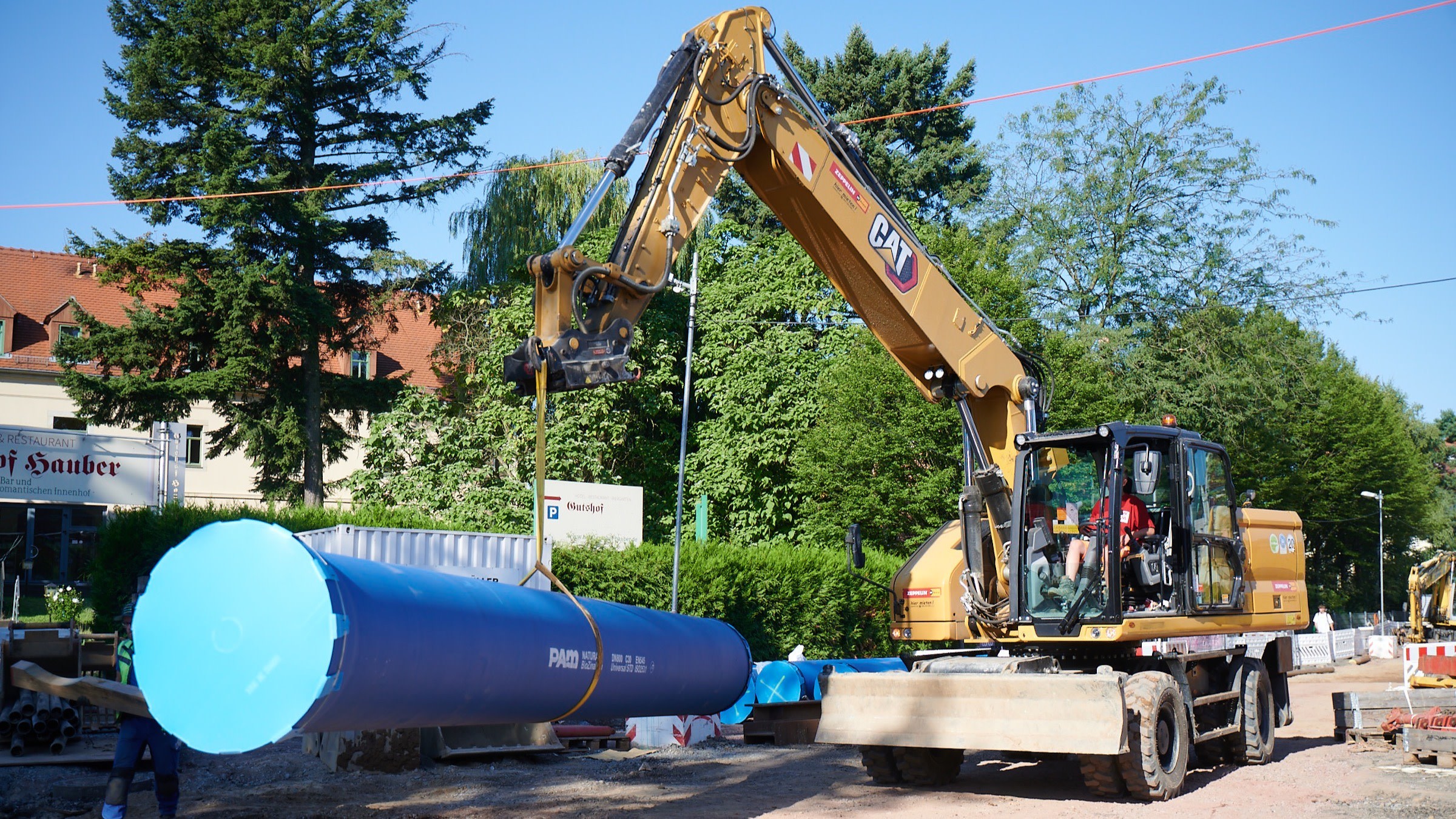 Ein gelber Bagger hebt ein langes, blaues Rohr. Es ist mit gelben Gurten am Baggerarm befestigt. Im Vordergrund ist ein Arbeiter zu sehen, der das Rohr führt. Die Baustelle ist von Bäumen und Gebäuden mit roten Dächern umgeben. Rechts im Bild sind weitere Baustellenmaterialien sowie ein Teil eines anderen Baggers erkennbar.