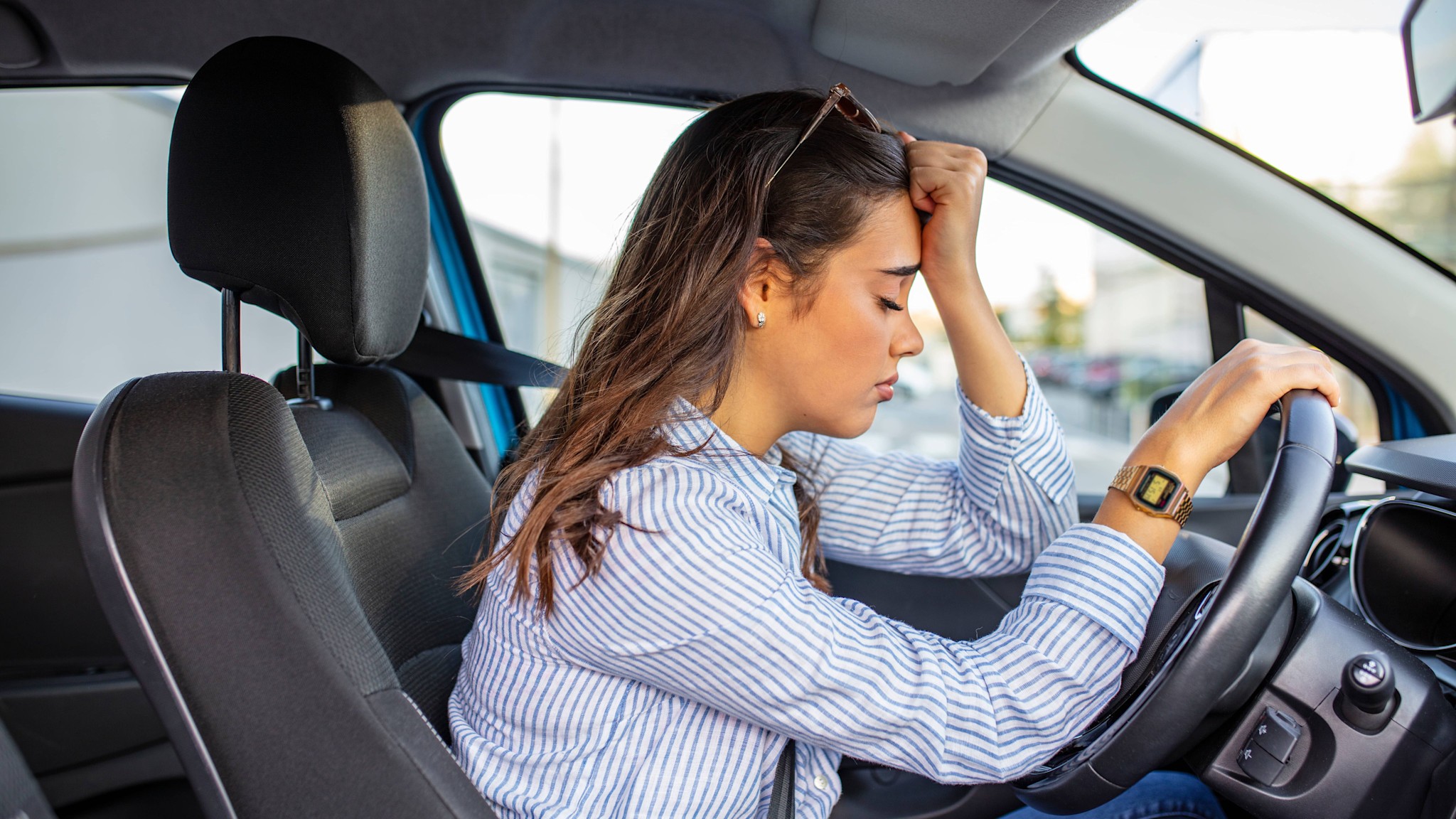 Fahrangst Header 16:9 Zu sehen ist eine Frau, die angegurtet in einem stehenden Auto sitzt. Sie lehnt gegen das Lenkrad und hält sich mit der linken Hand die Stirn.