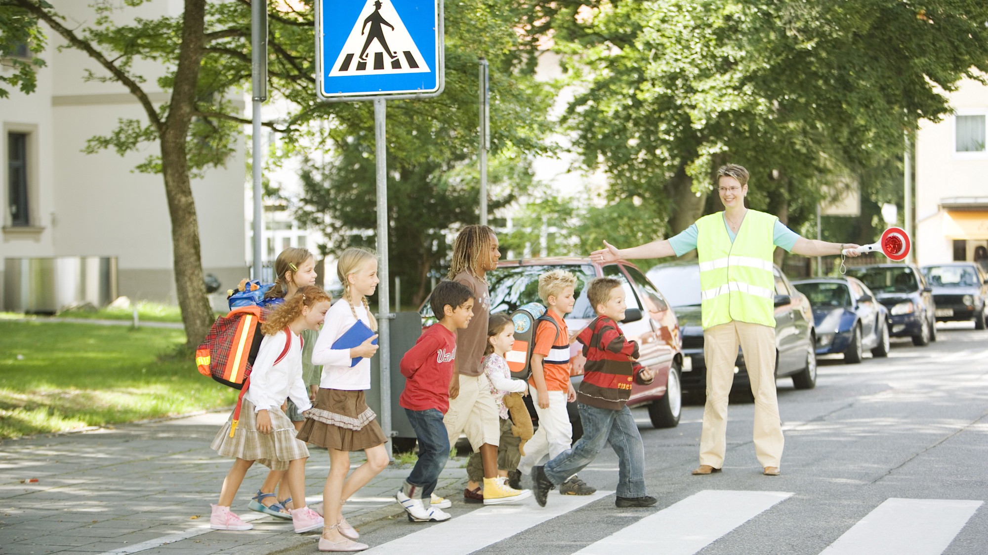 Kinder überqueren gemeinsam mit einer Verkehrshelferin einen Zebrastreifen in einer von Bäumen gesäumten Straße.