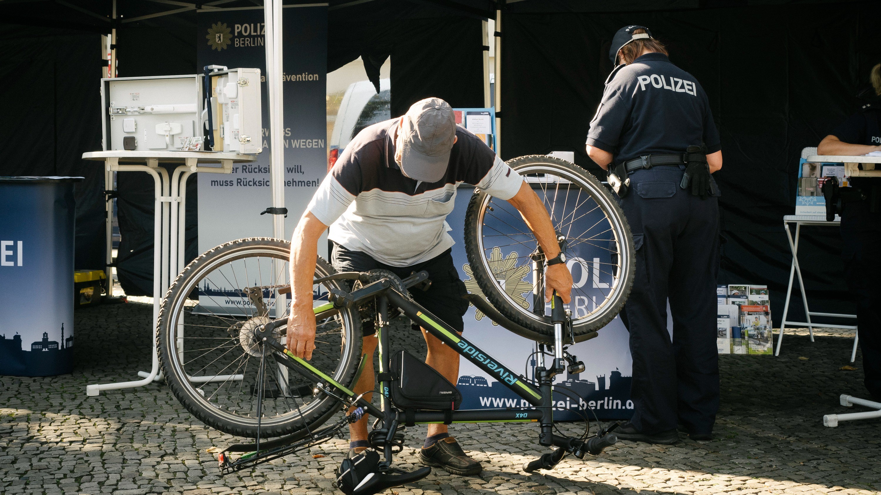 Ein älterer Herr liest die Rahmennummer von seinem umgedrehten Fahrrad ab. Im Hintergrund ist ein Aktionsstand mit der Aufschrift „Polizei Berlin“ zu sehen.