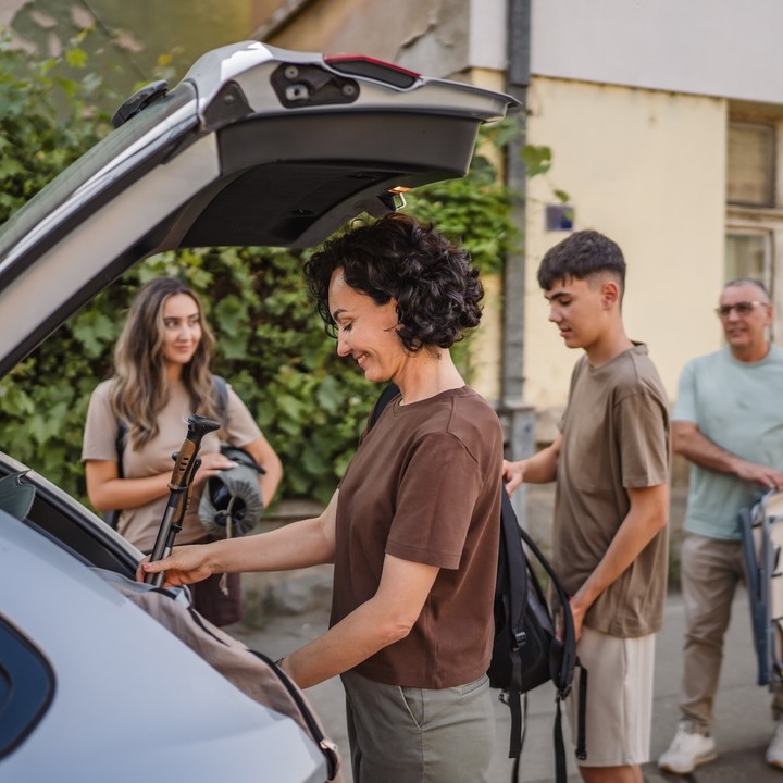 Eine Familie steht an der offenen Heckklappe eines Pkw. Sie beladen das Fahrzeug für die Urlaubsreise.