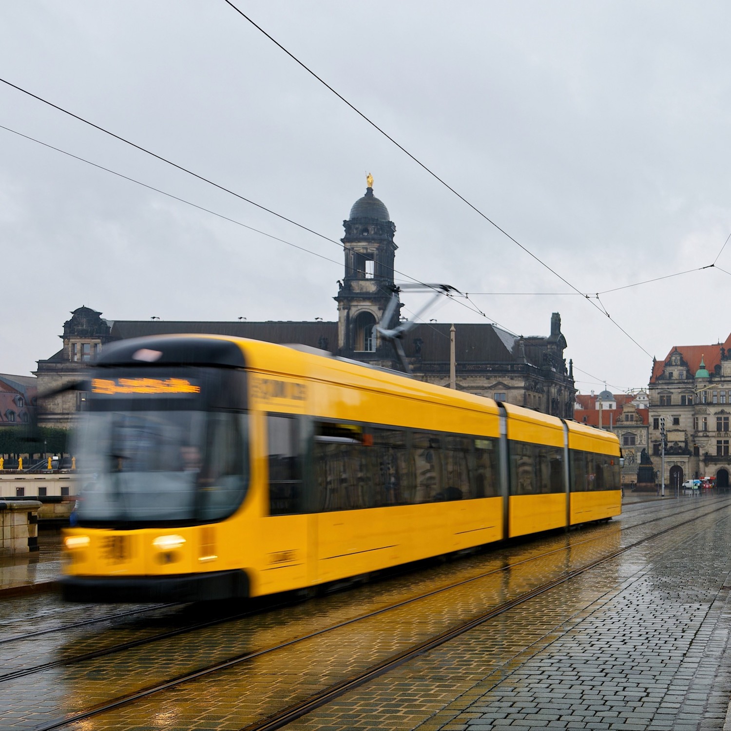 Header Straßenbahn 1:1 Eine gelbe Straßenbahn fährt im Regen eine gepflasterte Straße entlang. Im Hintergrund ist die Silhouette der Dresdner Altstadt zu sehen.
