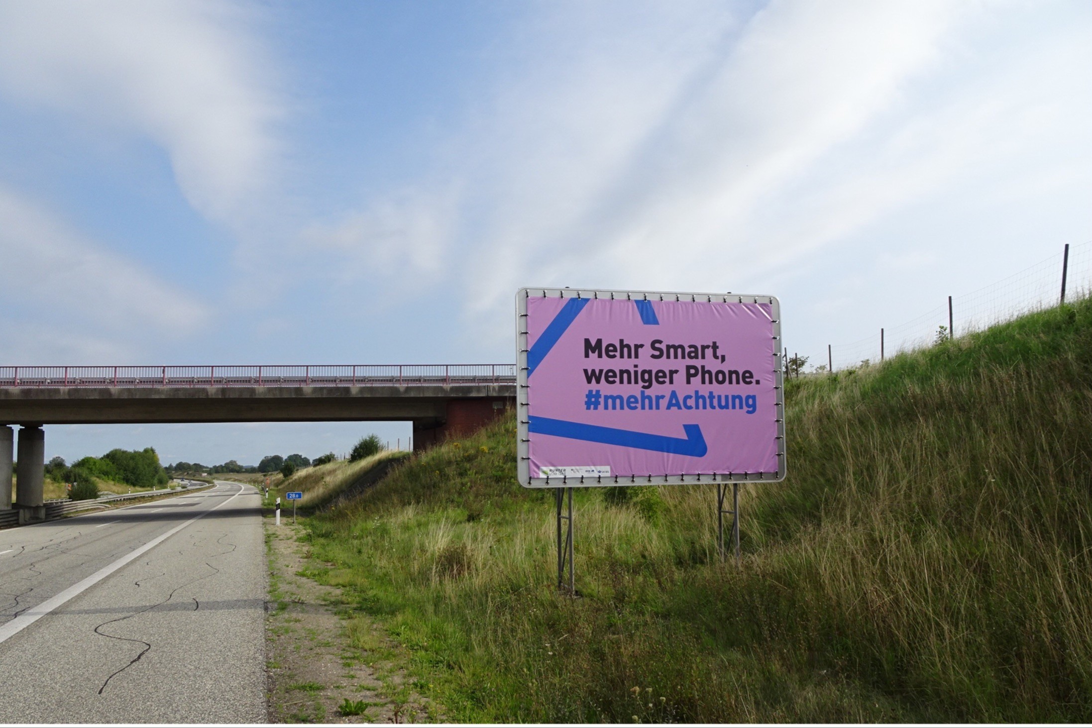 Neben einer Autobahn steht ein großer Plakataufsteller mit einem Autobahnplakat. Darauf ist Folgendes zu sehen: Vor einem blauen Dreieck auf pinkfarbenem Hintergrund, das an das Verkehrszeichen „Gefahrenstelle“ erinnert, steht der Text: „Mehr Smart, weniger Phone. Mehr Achtung.“ Im Hintergrund sieht man eine Autobahnbrücke.