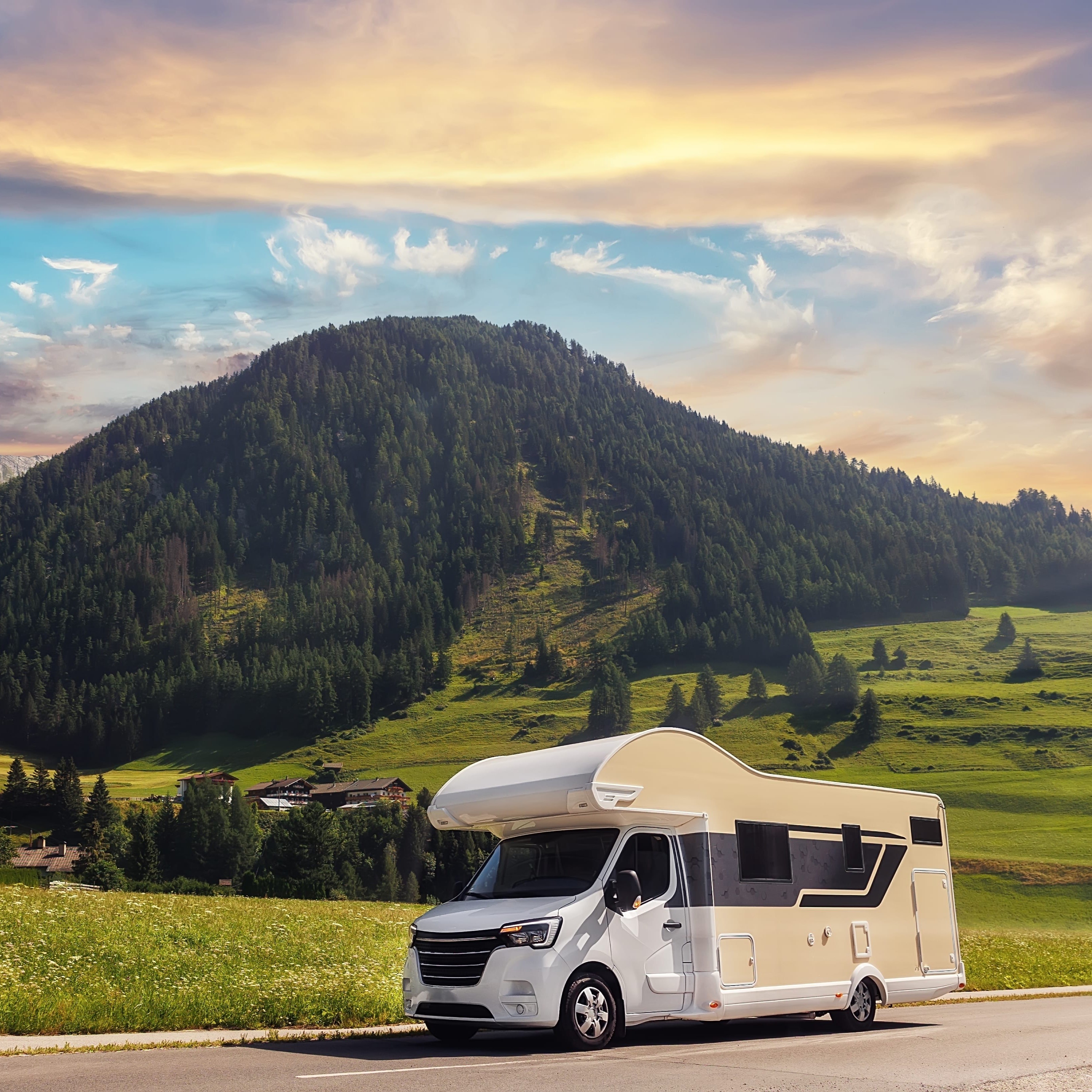 Wohnmobilurlaub – Wohnmobil am Straßenrand 1:1 Ein modernes, weiß-beiges Wohnmobil steht am Straßenrand vor einer malerischen Berglandschaft mit grünen Wiesen und bewaldeten Hügeln bei Sonnenuntergang. Das Bild symbolisiert den Urlaub mit dem Wohnmobil.