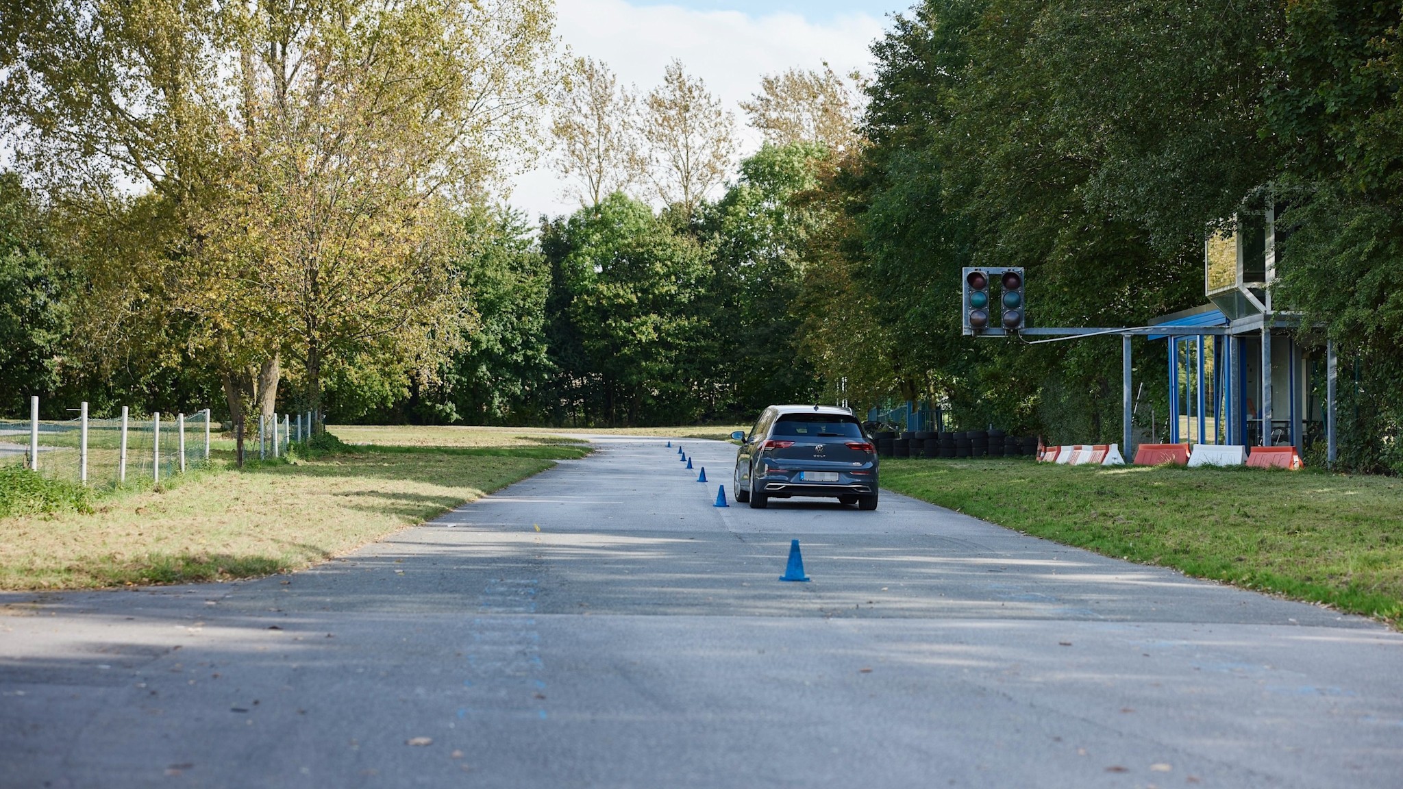 Fahrsicherheitstraining Slalom Ein Auto fährt auf einer Straße mit blauen Verkehrskegeln, die eine Spur markieren. Rechts ist eine ausgeschaltete Ampel zu sehen. Die Umgebung ist von Bäumen gesäumt.