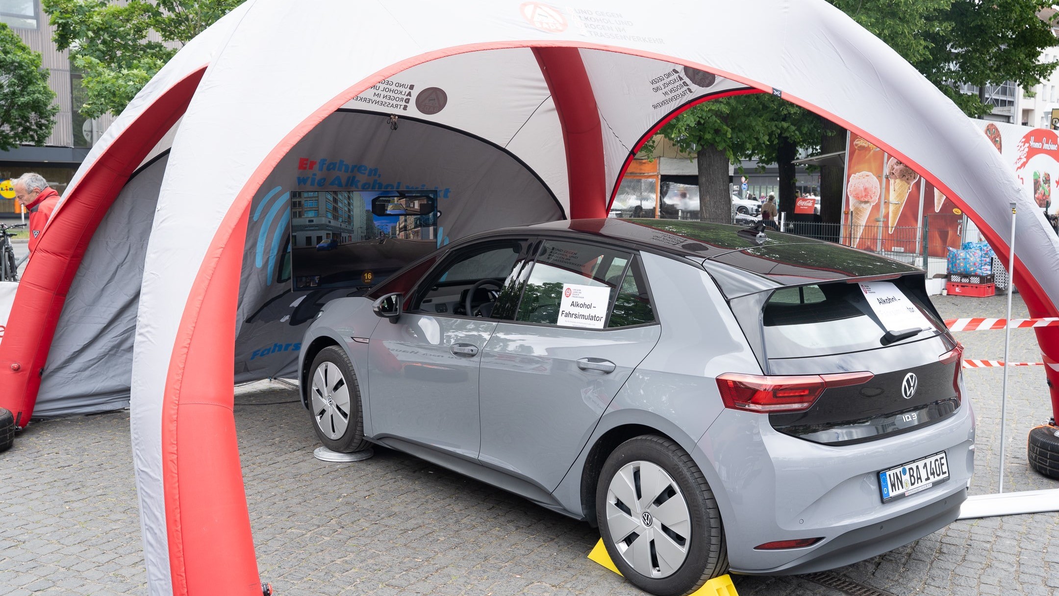 Ein Auto mit dem Schild „Alkohol-Fahrsimulator“ steht unter einem grauen Kuppelpavillon mit roten Streben. Das Fahrzeug ist zur Stabilisierung auf gelben Keilen abgestellt. Die mobile Verkehrssicherheitsstation befindet sich im Freien – im Hintergrund sind Bäume und Gebäude sichtbar.