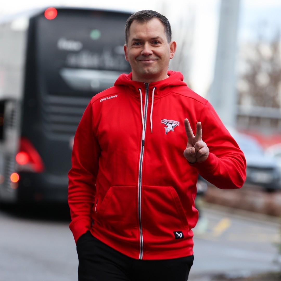 Busfahrer Enrico Bernig trägt einen roten „Kölner Haie“-Hoodie und zeigt das Peace-Zeichen. Im Hintergrund ist ein Bus zu sehen.