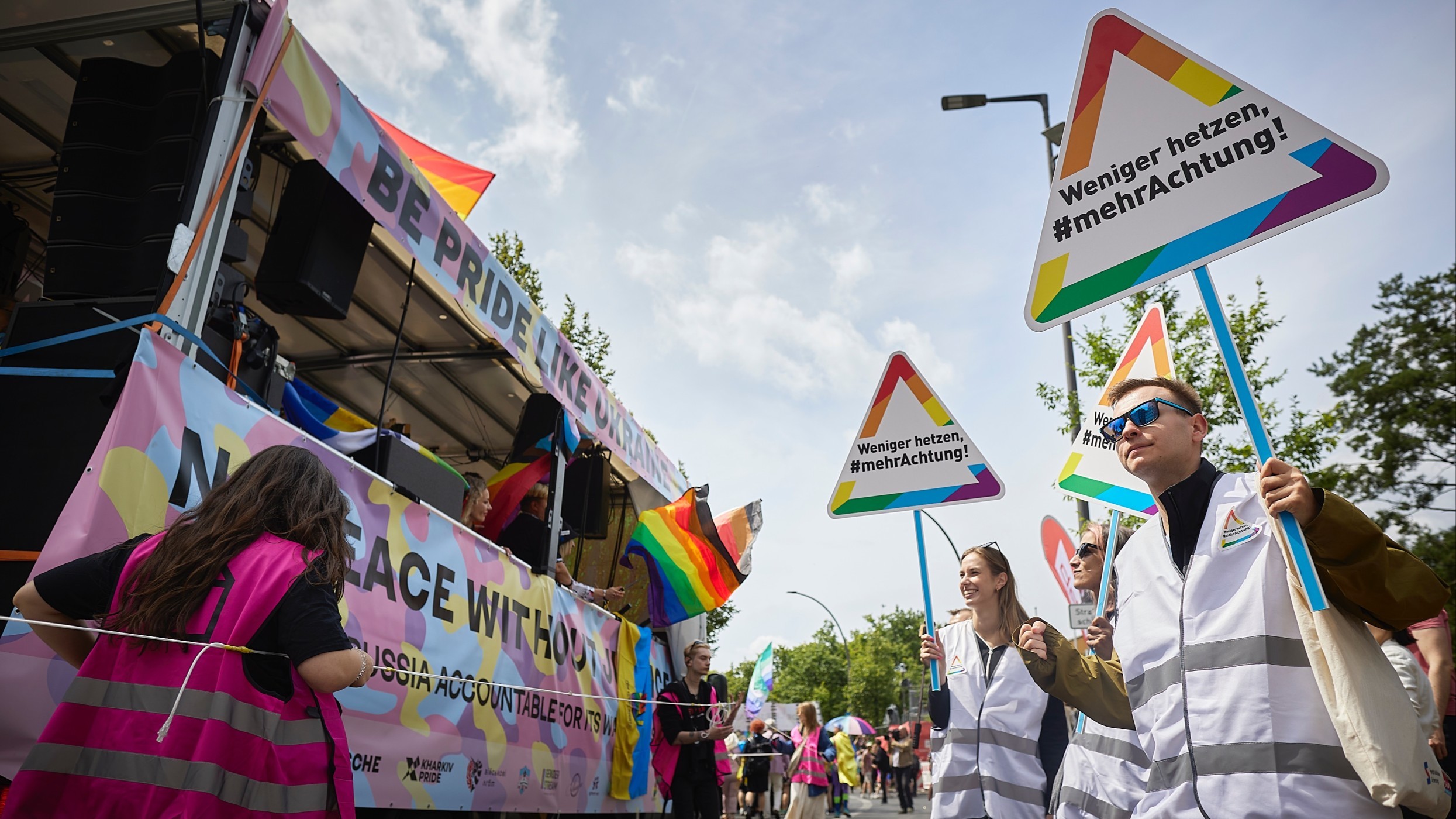 Zwei Frauen und ein Mann stehen an einer Straße und halten Schilder in der Hand. Darauf steht: „Weniger hetzen, mehr Achtung!“ Links von ihnen fährt ein geschmückter CSD-Wagen an ihnen vorbei. 