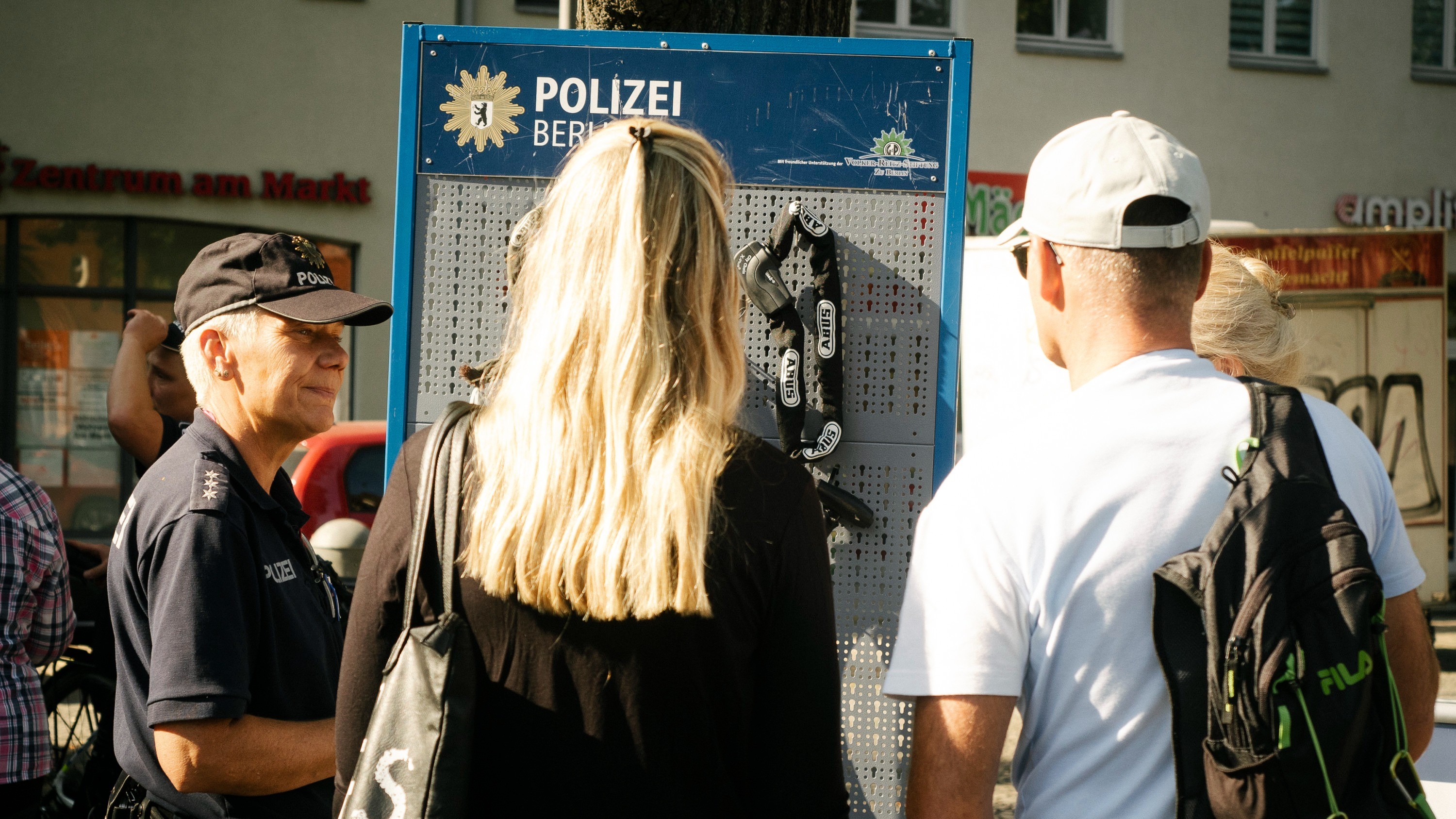 Eine Polizistin zeigt einer Frau mit blondem Haar und einem Mann mit weißem T-Shirt und weißer Kappe verschiedene Fahrradschlösser.