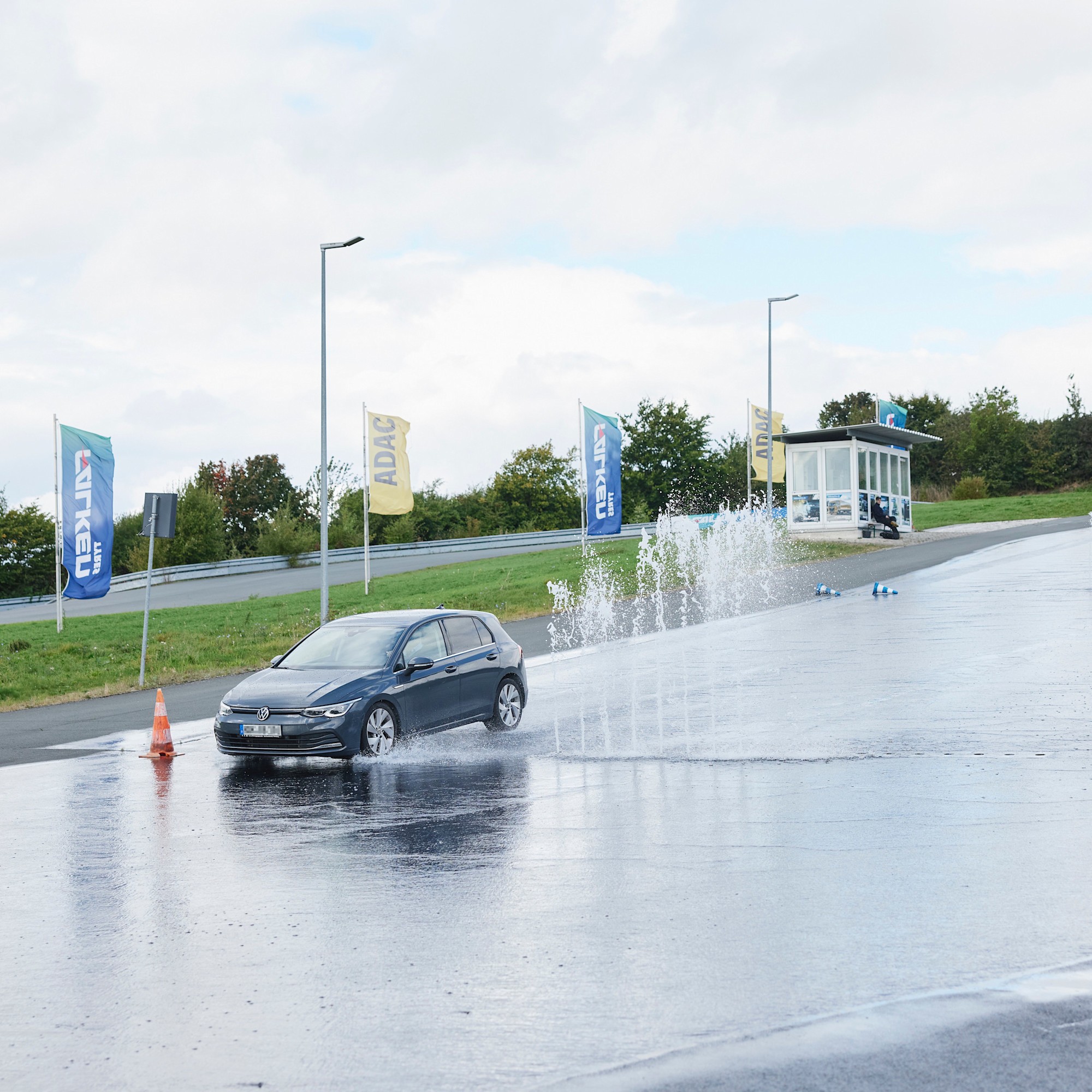 Fahrsicherheitstraining Nasse Fahrbahn Ein Auto fährt über eine nasse Fahrbahn in einem Fahrsicherheitszentrum. Wasserfontänen spritzen auf, während das Fahrzeug vorbeifährt. Im Hintergrund sind Flaggen und ein kleines Gebäude zu sehen.