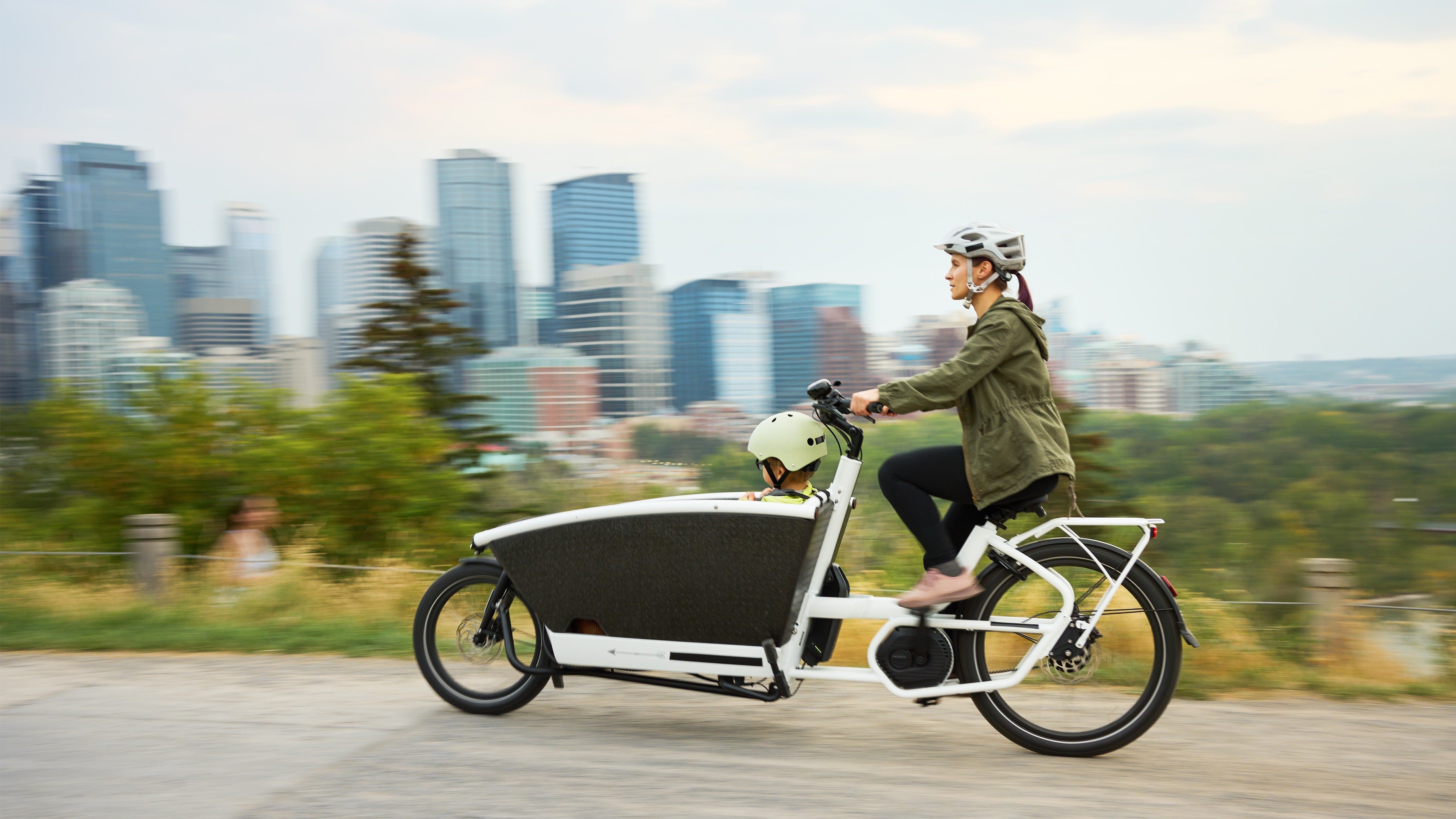 Header Lastenrad 16:9 Eine Frau mit Helm fährt ein Lastenrad mit Transportbox, in der ein Kind mit grünem Helm sitzt. Sie fahren auf einem Weg vor einer grünen Parklandschaft und Hochhäusern.