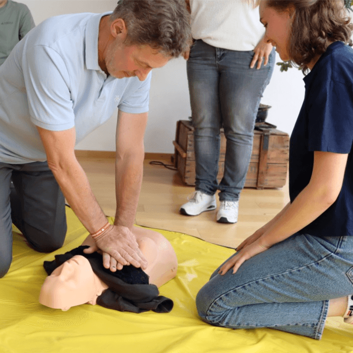 mA Erste Hilfe Kurs Björn Steiger Stiftung Mehrere Personen nehmen an einem Erste-Hilfe-Kurs teil. Ein Mann kniet auf dem Boden und führt eine Herzdruckmassage an einer Übungspuppe durch.