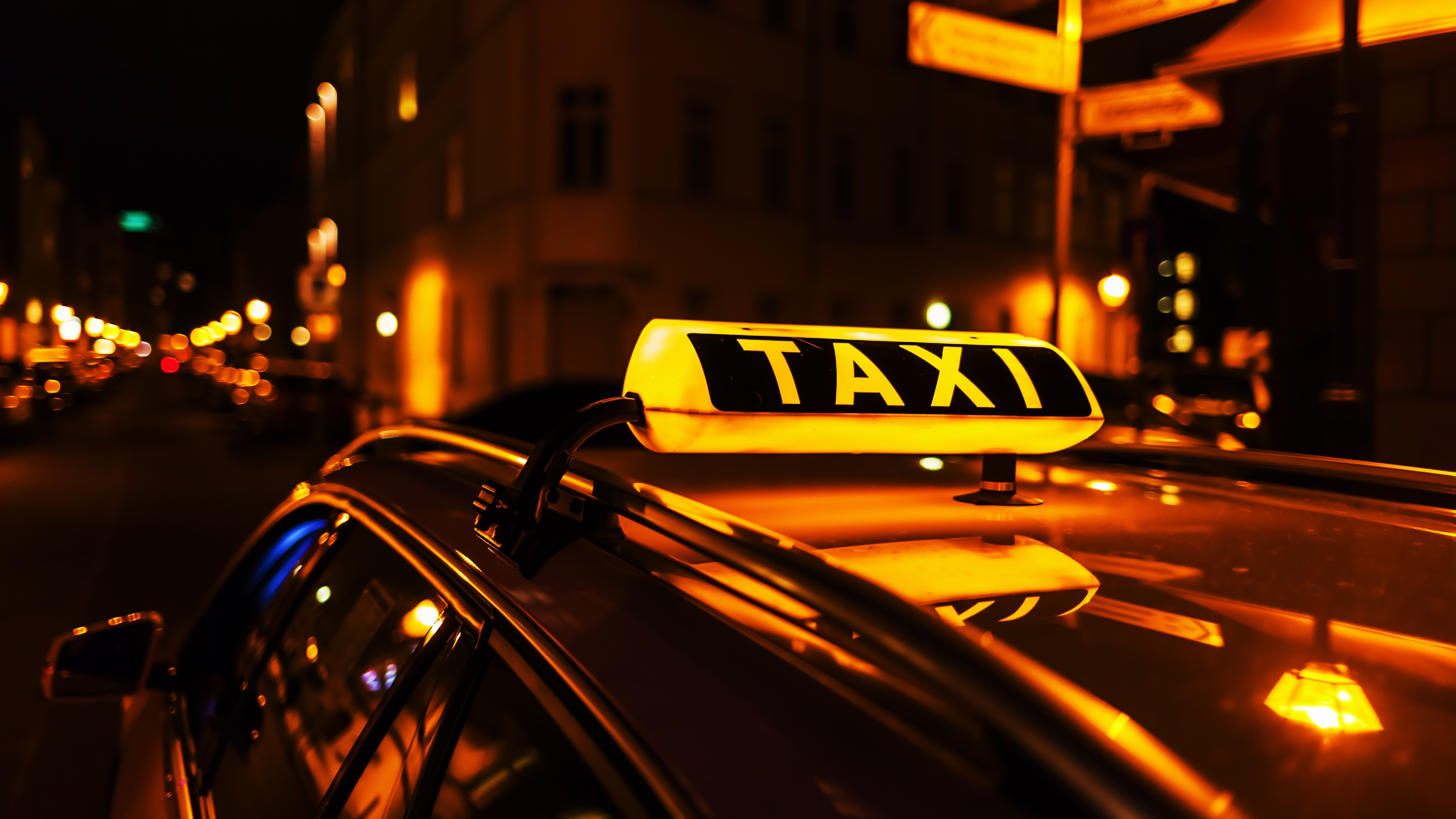 Jugendtaxi: Bild Taxi-Schild 16:9 Ein Taxi-Schild leuchtet bei Nacht in gelber Farbe auf dem Deck eines Autos. Im Hintergrund sind verschwommen Häuser zu sehen.