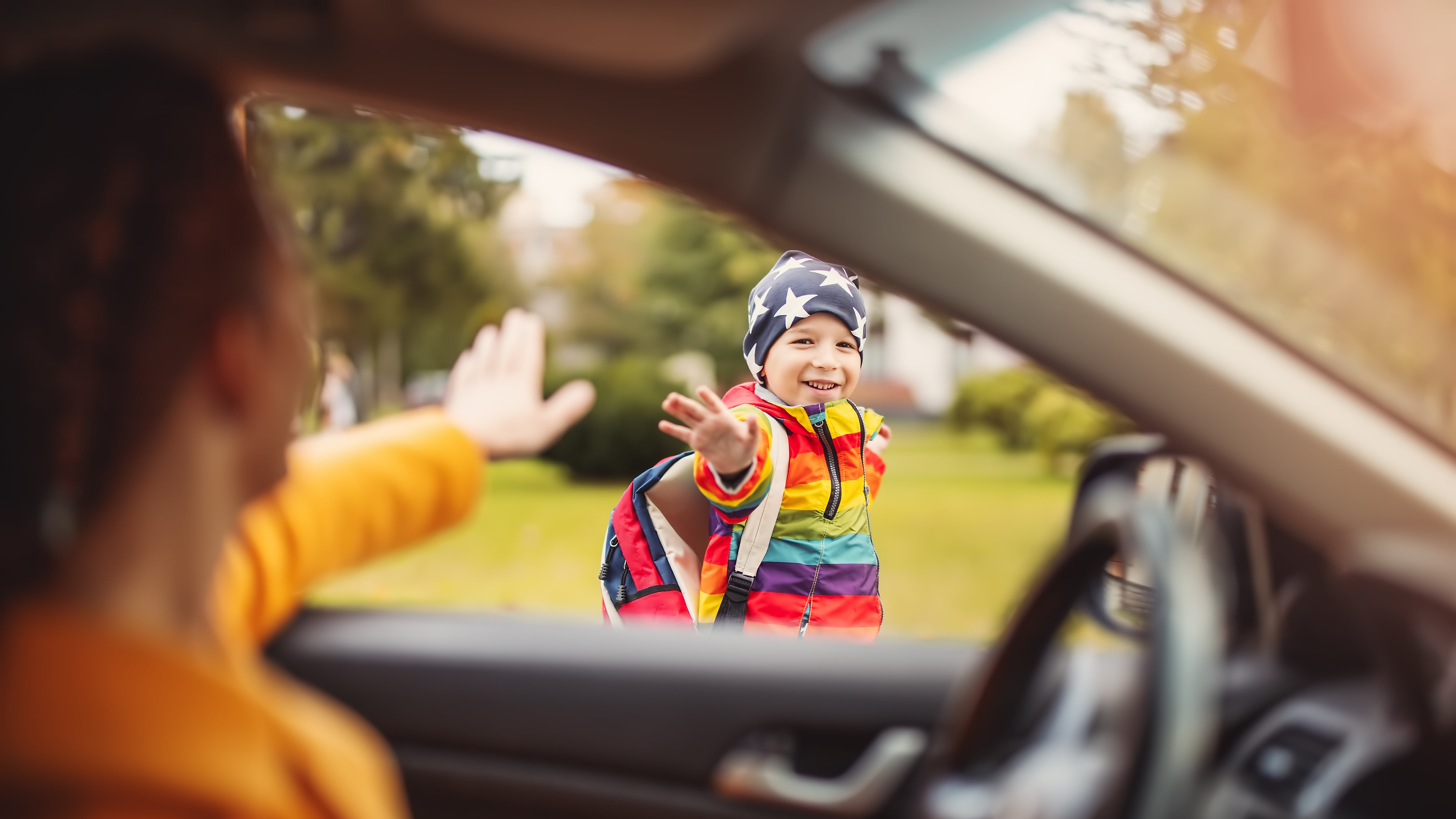 Perspektive des Inneren eines Autos. Eine Frau, deren Arm und Schulter im Vordergrund unscharf zu sehen sind, winkt mit der Hand nach draußen. Durch die offene Autotür oder das Fenster blickt ein lächelndes Kind mit einem bunten Schulranzen und einer Mütze mit Sternenmuster. Das Kind winkt ebenfalls zurück.