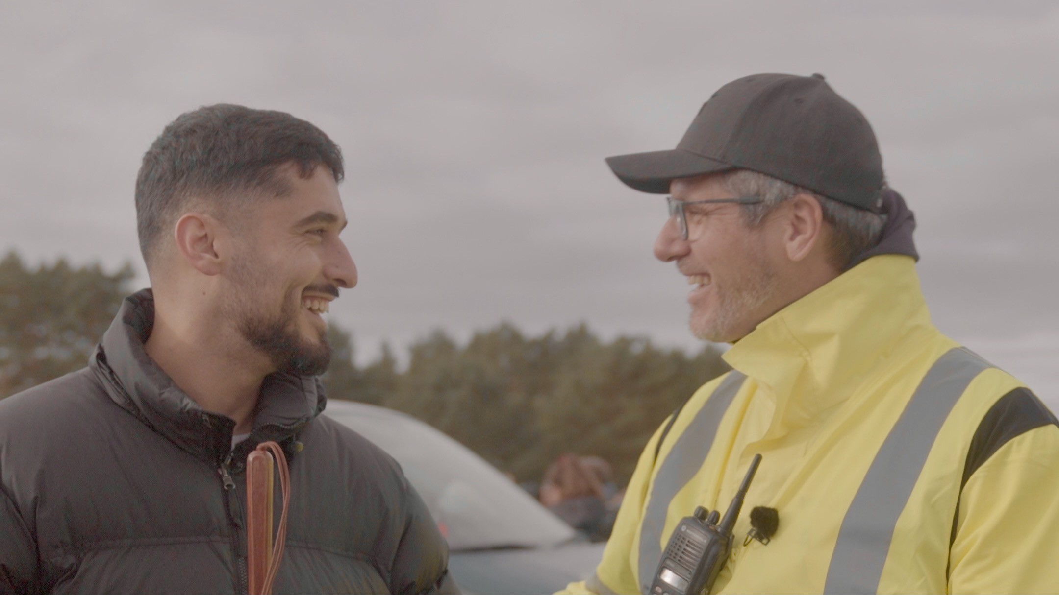 Yusuf Alp Ay mit schwarzer Jacke und Blindenlangstock und Stephan Ackerschewski mit gelber Warnjacke und Funkgerät stehen sich im Freien gegenüber.